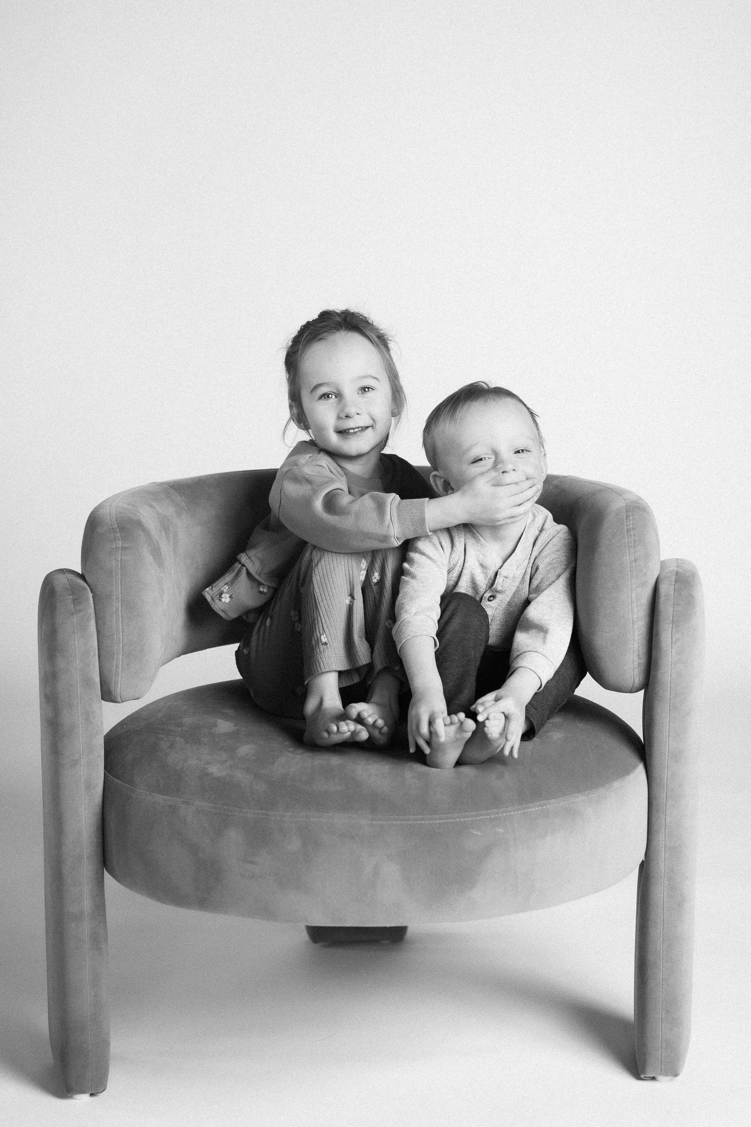 Black and white photo series showing two babies lounging together on a curved modern chair in playful poses.