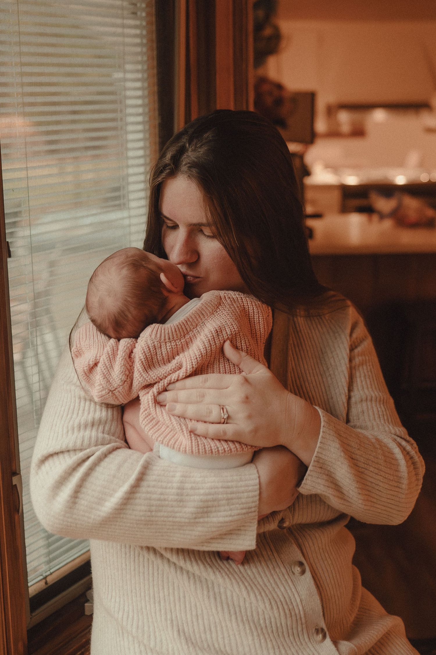 A tender moment between parent and baby in warm lighting as they embrace by a window with venetian blinds.
