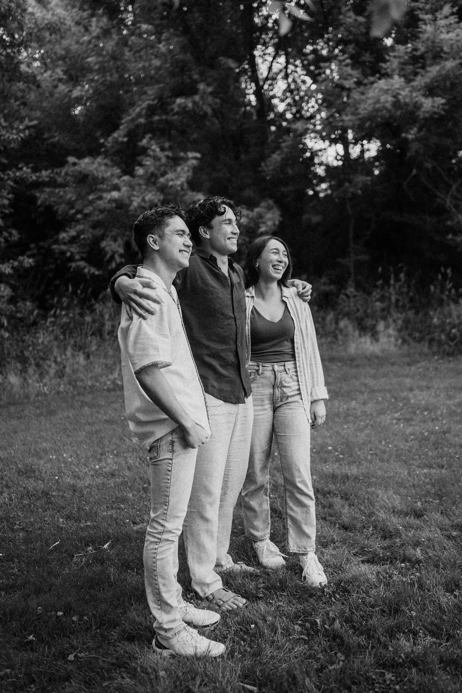 Three smiling friends embrace outdoors in a black and white photo on a grassy field with trees behind them.