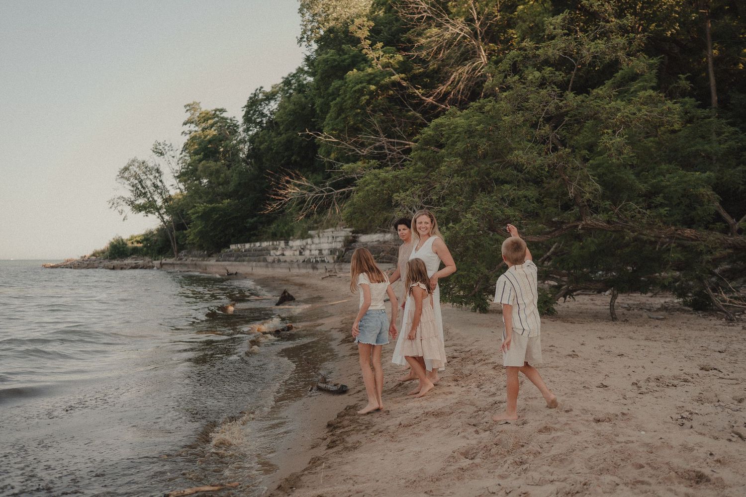 Family with children walking along a sandy beach shore surrounded by lush green trees at golden hour.