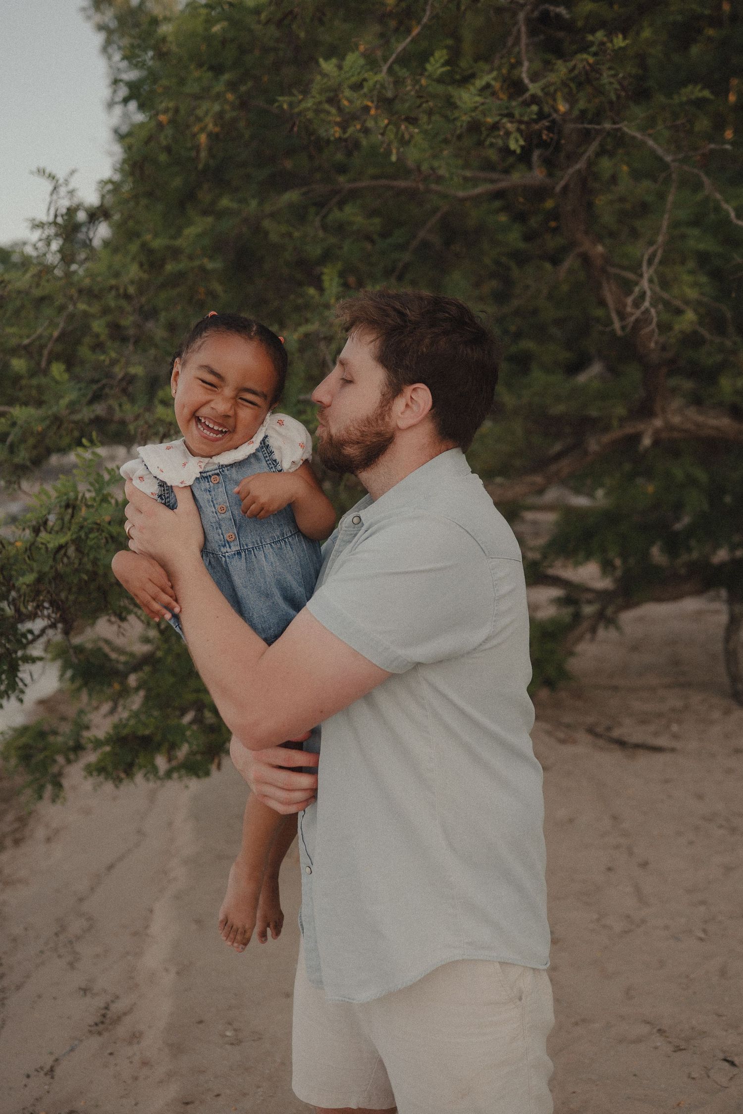 A bearded man lifts and kisses a giggling toddler girl in denim overalls on a sandy beach.