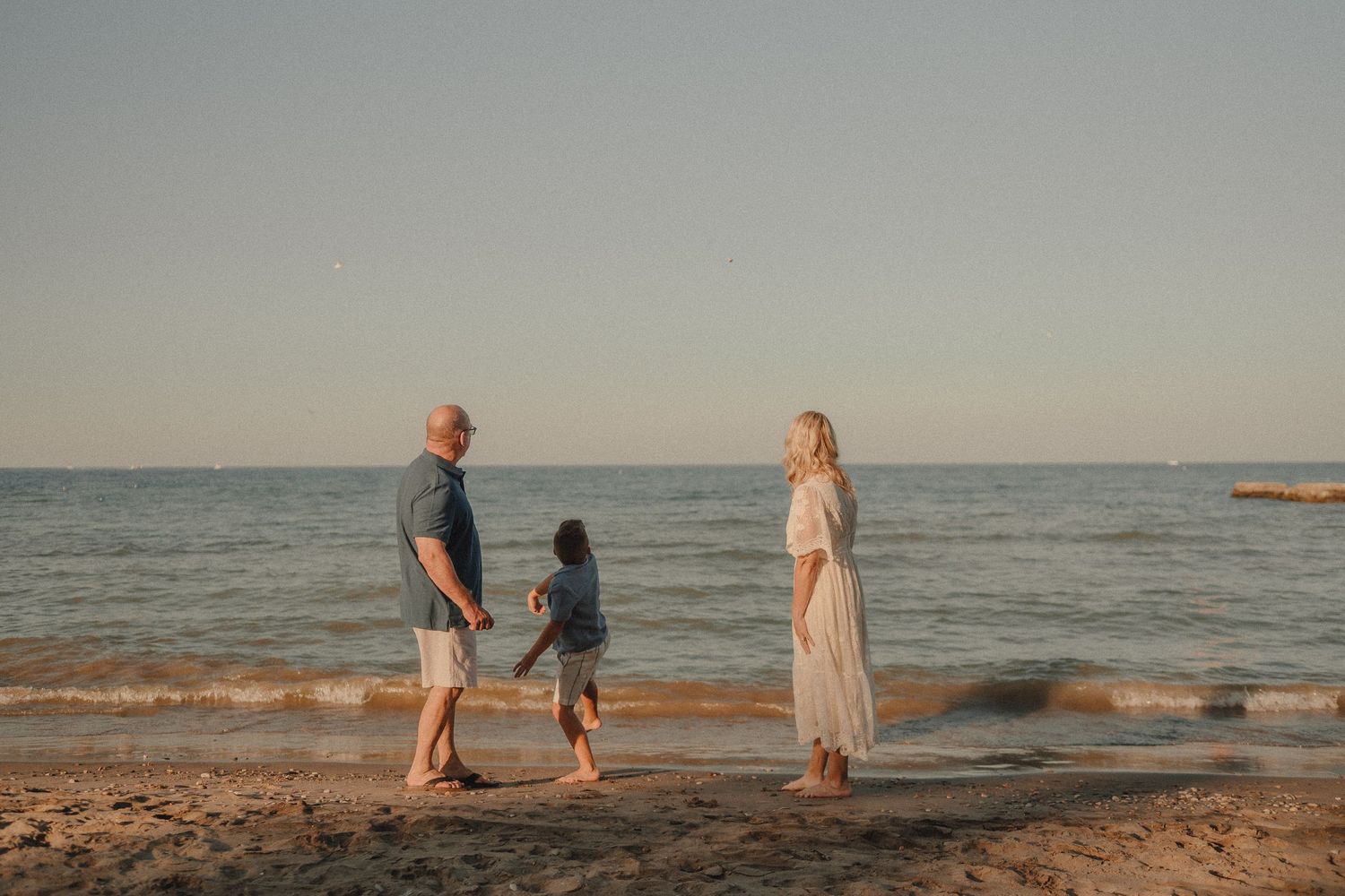 Family of three standing together on sandy beach at sunset with calm ocean behind them.