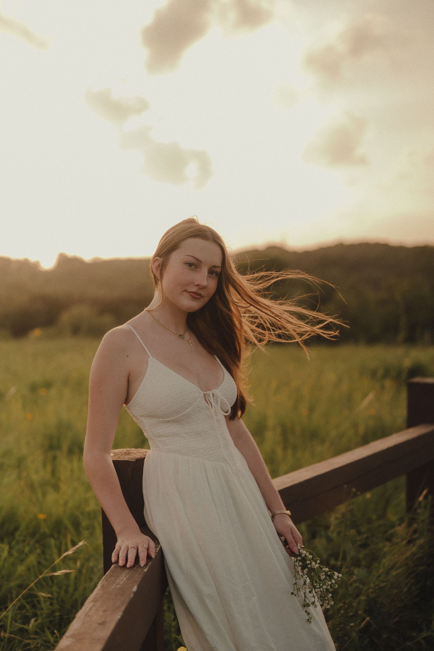 Young woman in white sundress leans on wooden fence in green meadow during golden sunset.