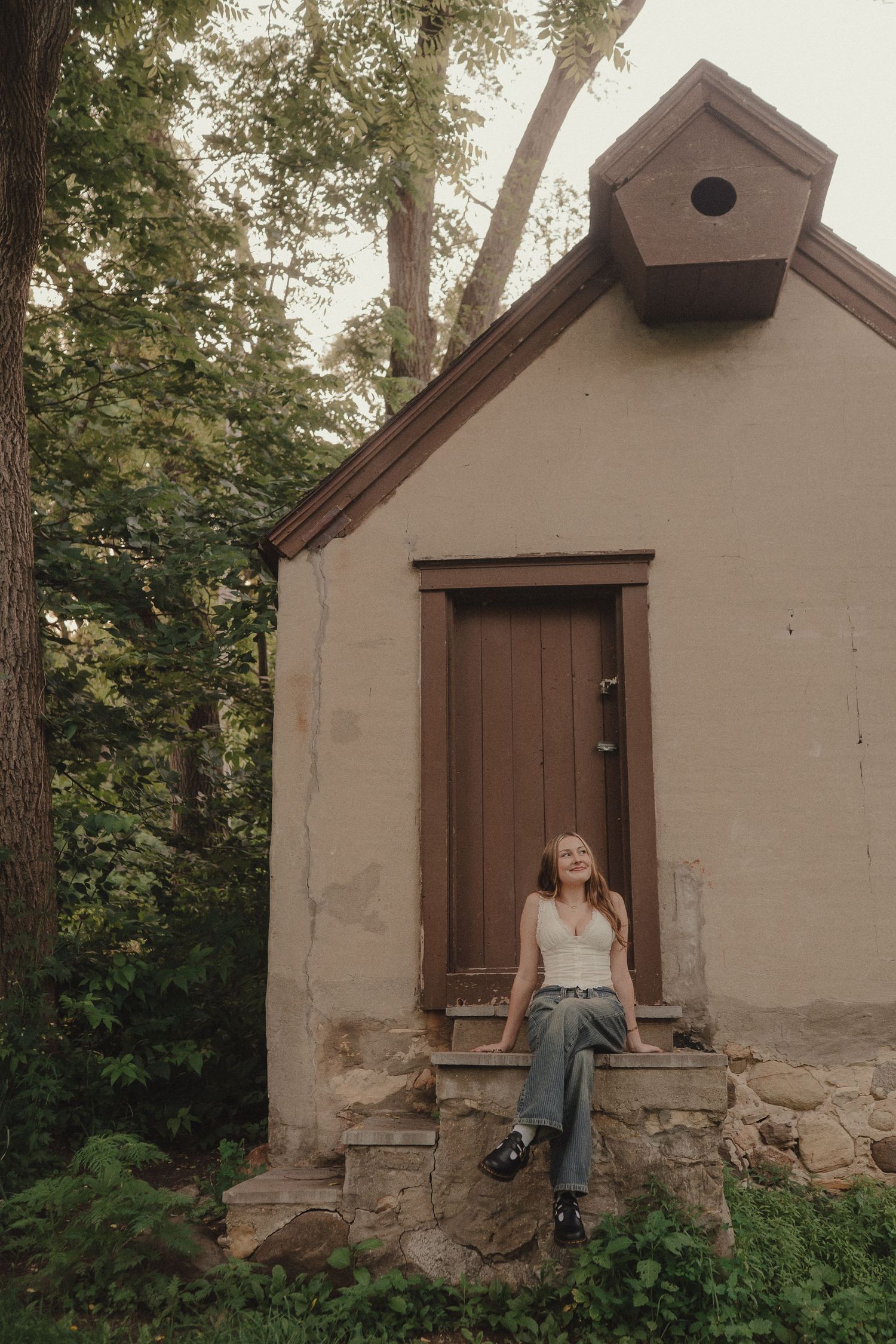 Young woman sitting on stone steps outside a rustic beige building surrounded by trees.