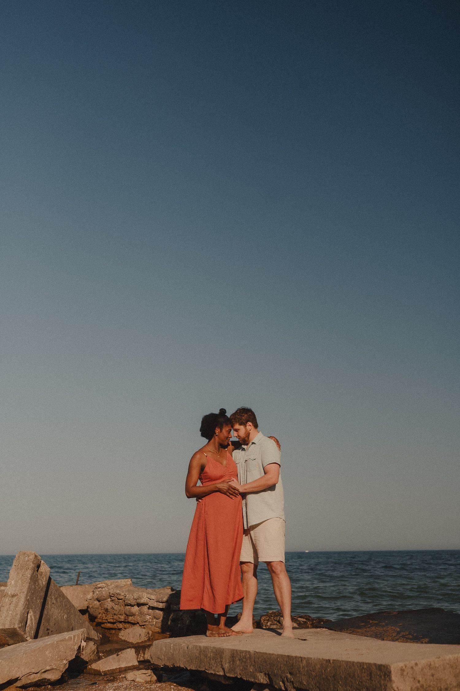Expecting interracial couple standing close together on rocky lakeside pier under clear blue sky at sunset.