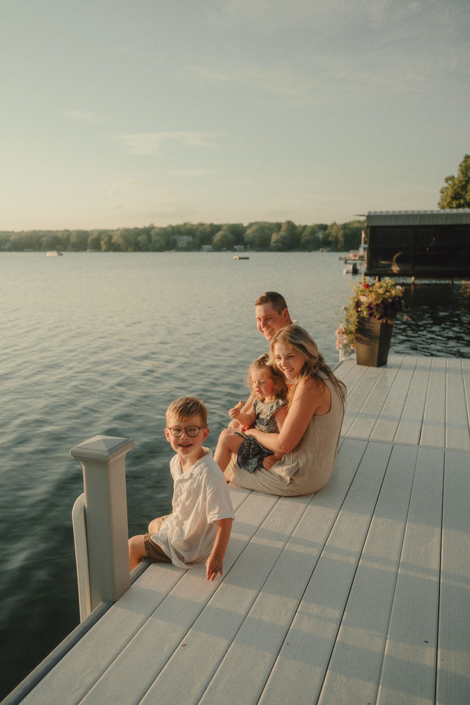 Family of four sitting on a white dock by a calm lake at golden hour sunset.