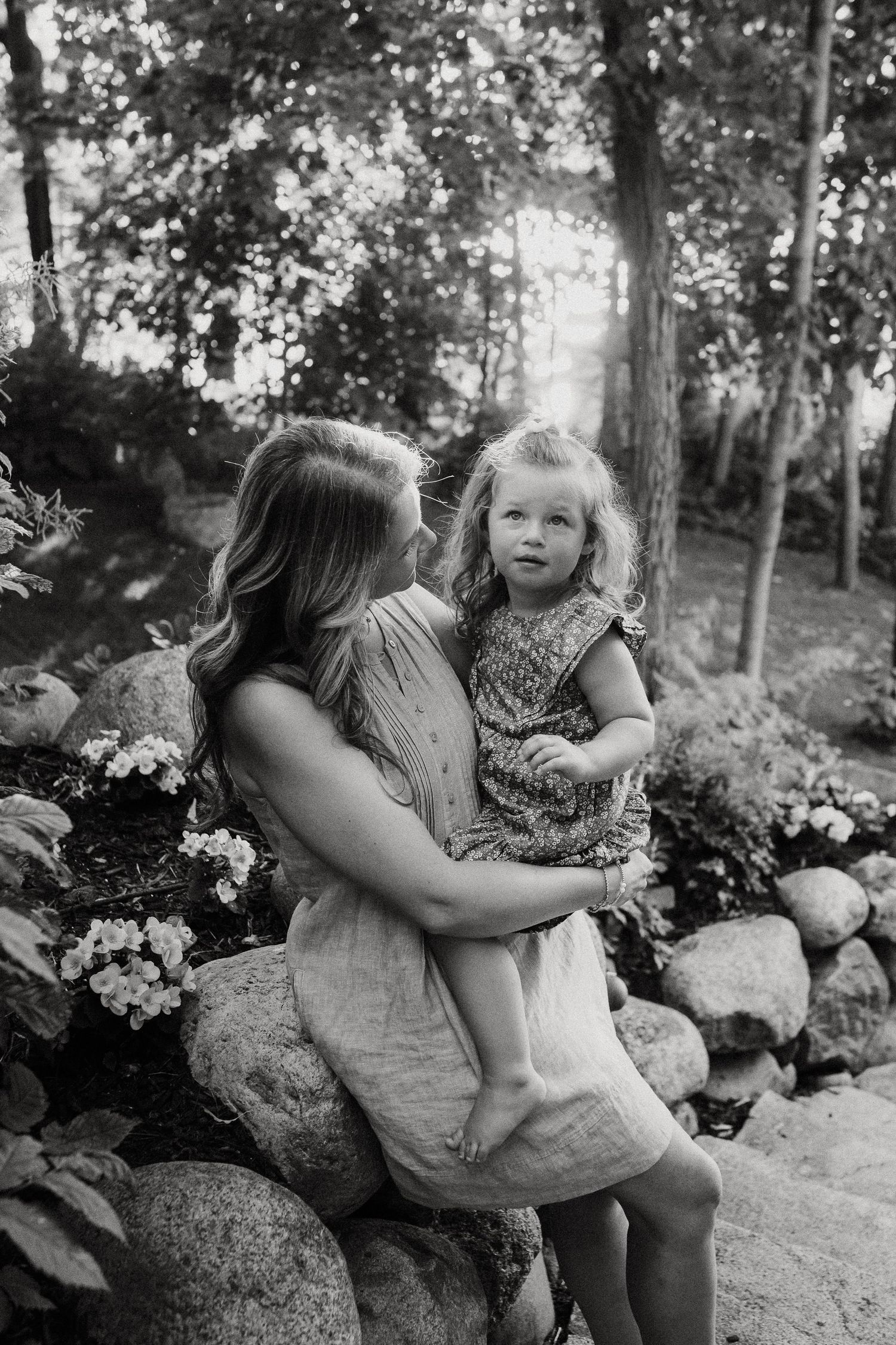 Black and white photo of a mother holding her young blonde daughter near a stone wall with flowers.