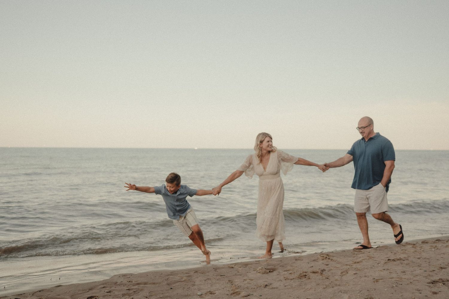 Family of three joyfully walking on a sandy beach at sunset, parents swinging child between them near calm ocean waters.