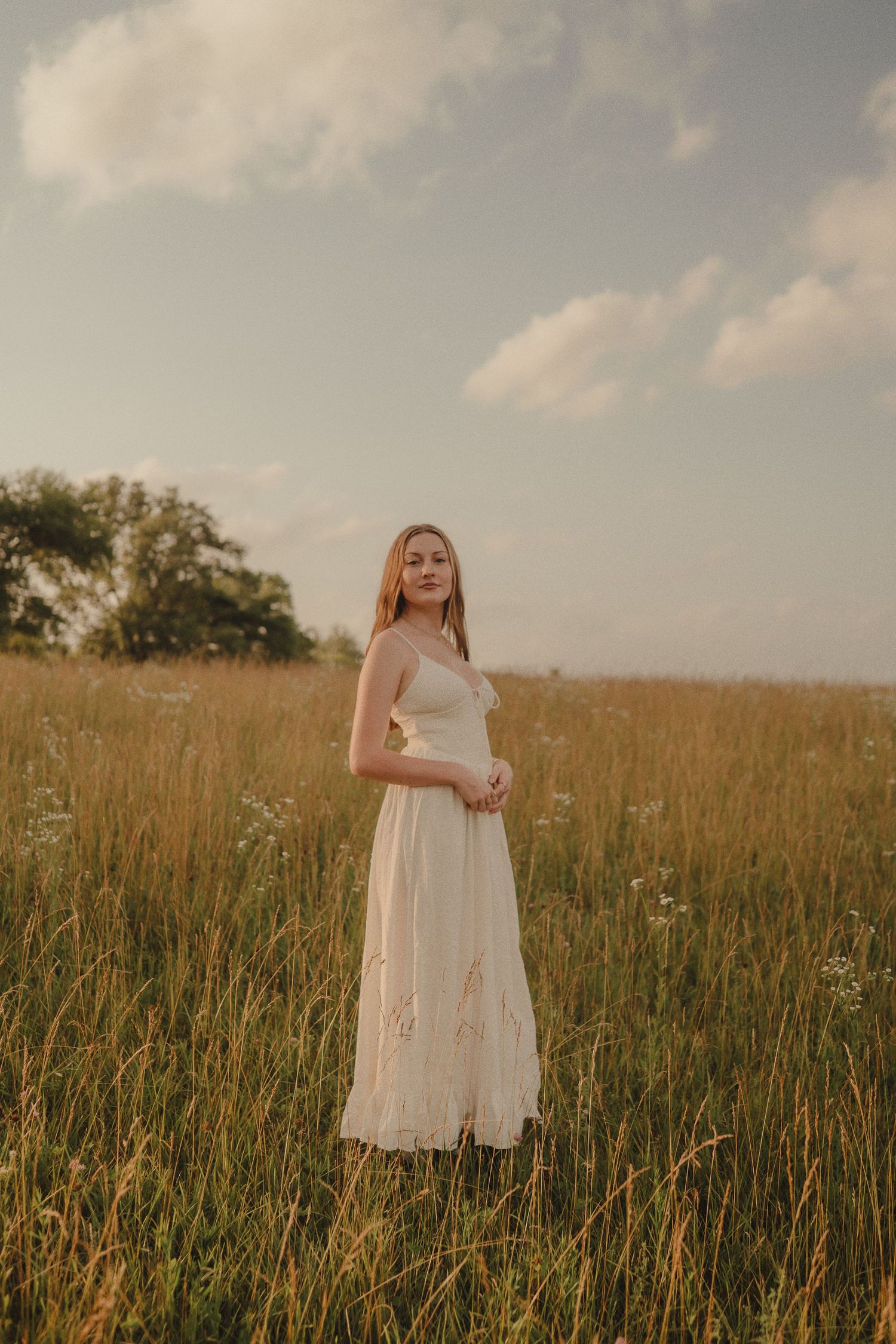 Woman in white sundress standing in tall golden meadow grass under cloudy sky.