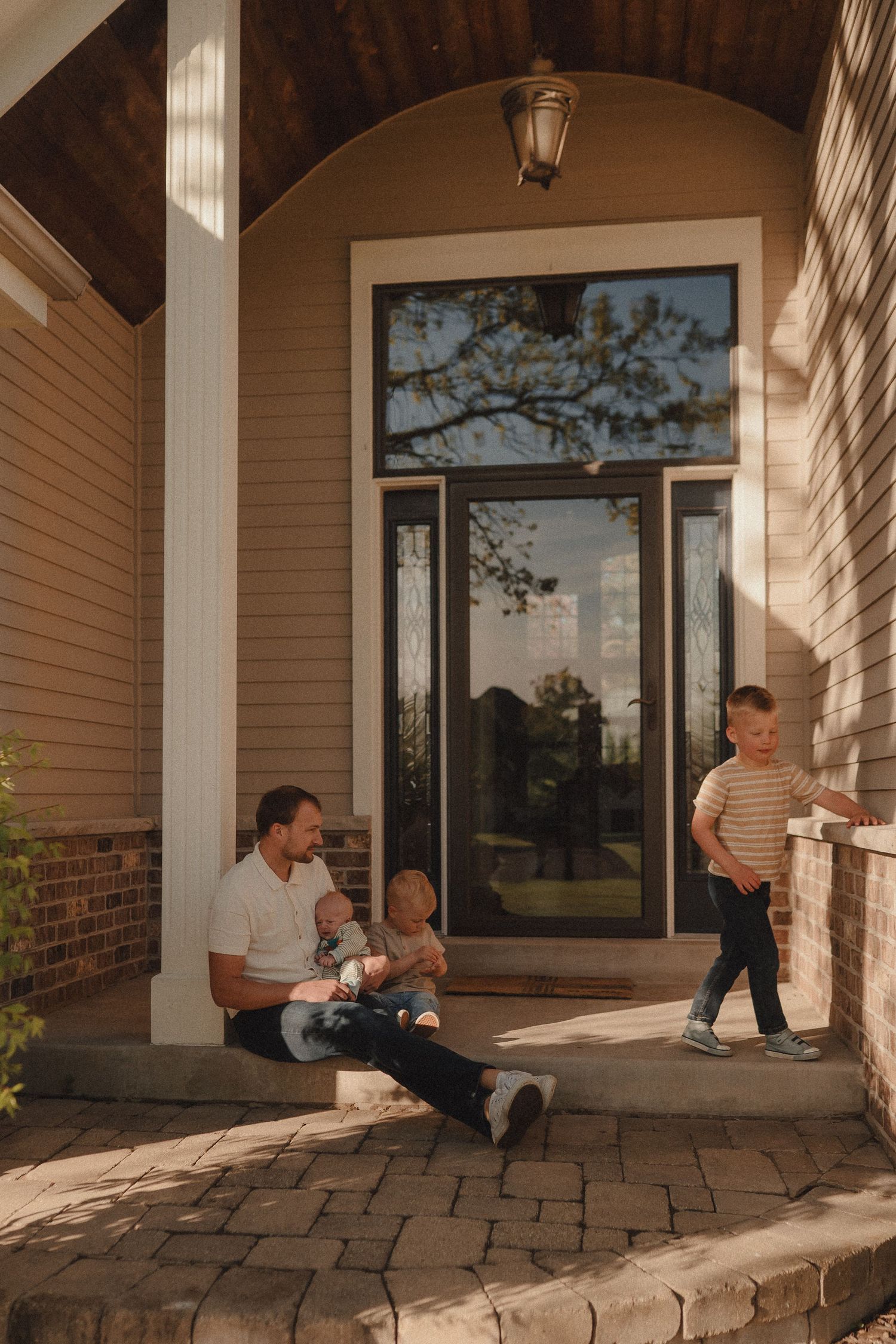 A father sits on a front porch holding an infant while a young boy plays on the steps nearby.