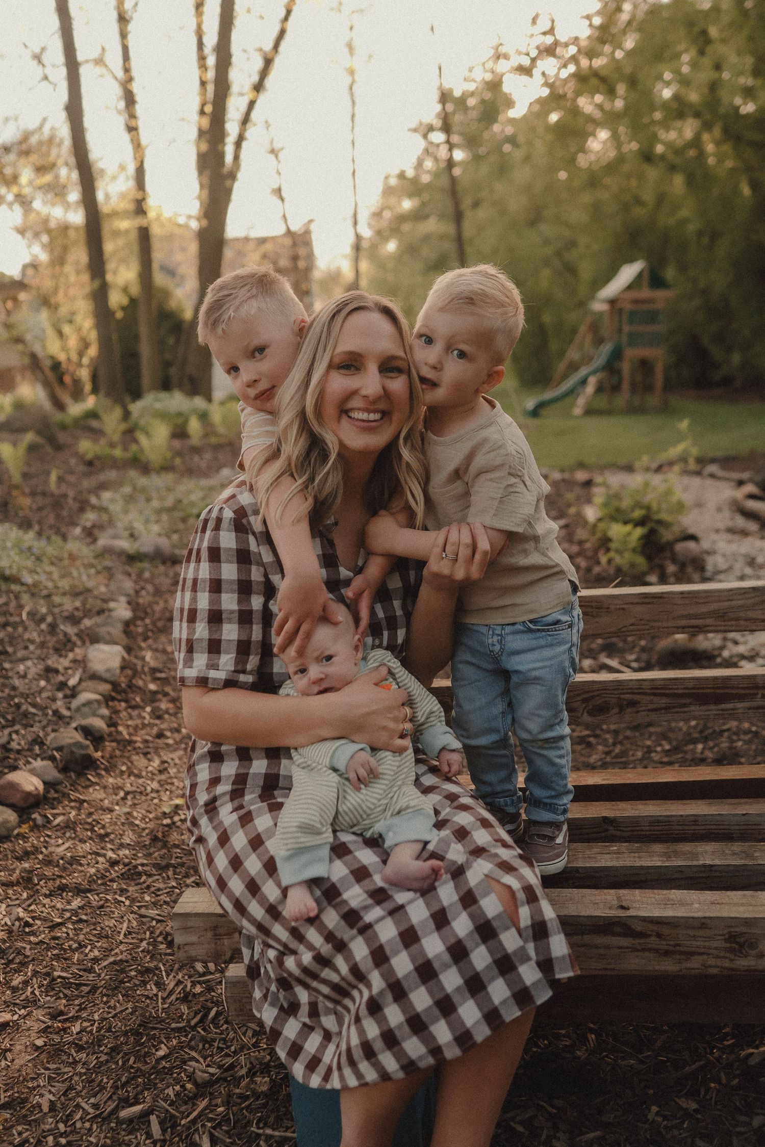 A joyful mother in a plaid dress sits outdoors holding her baby while two young boys embrace her warmly.