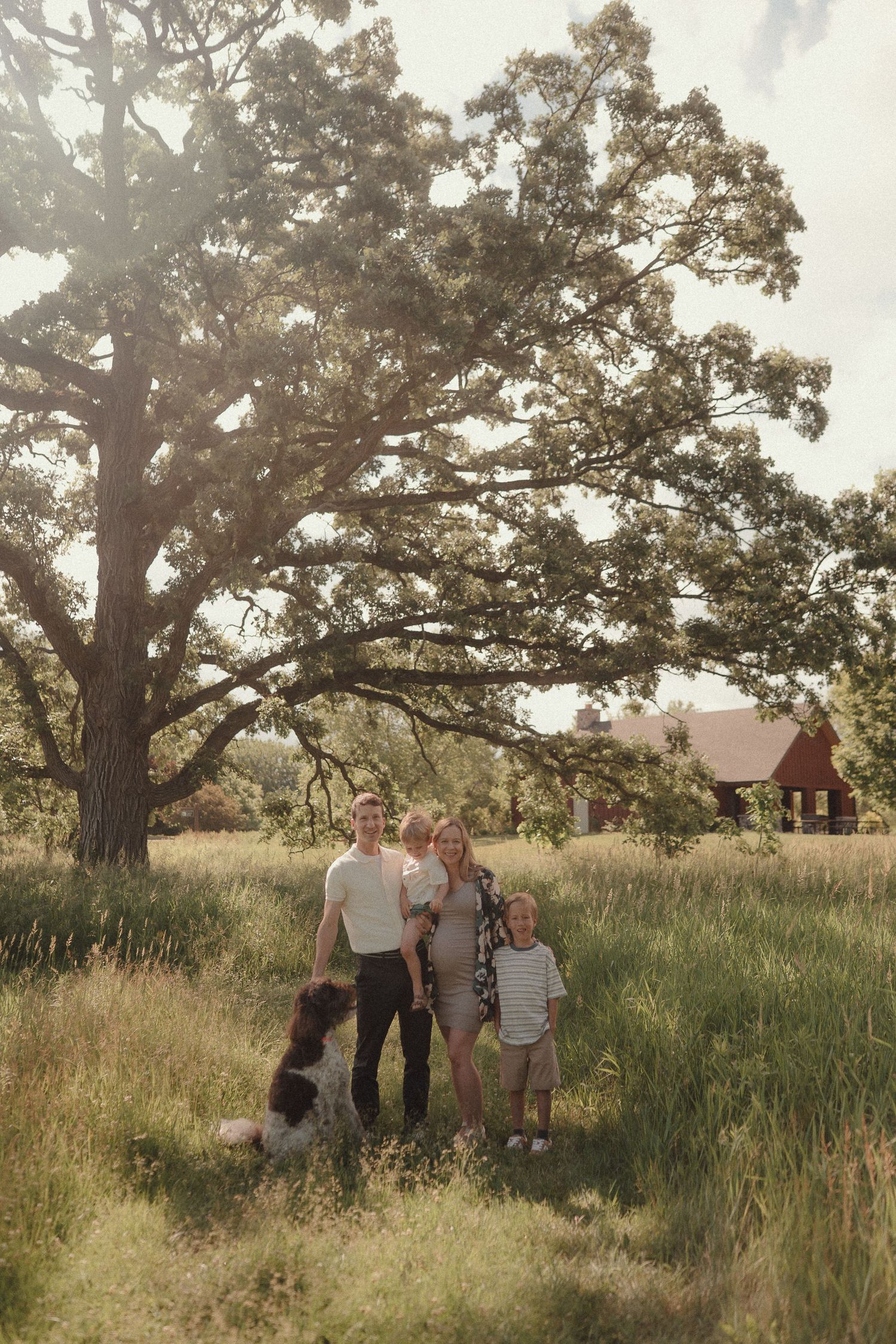 Family with dog posing in sunny meadow with large oak tree and red barn in background.