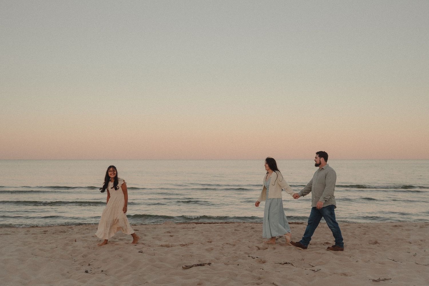 Three people walk on a sandy beach at sunset, two women in flowing dresses and a man in casual attire near calm ocean waves.