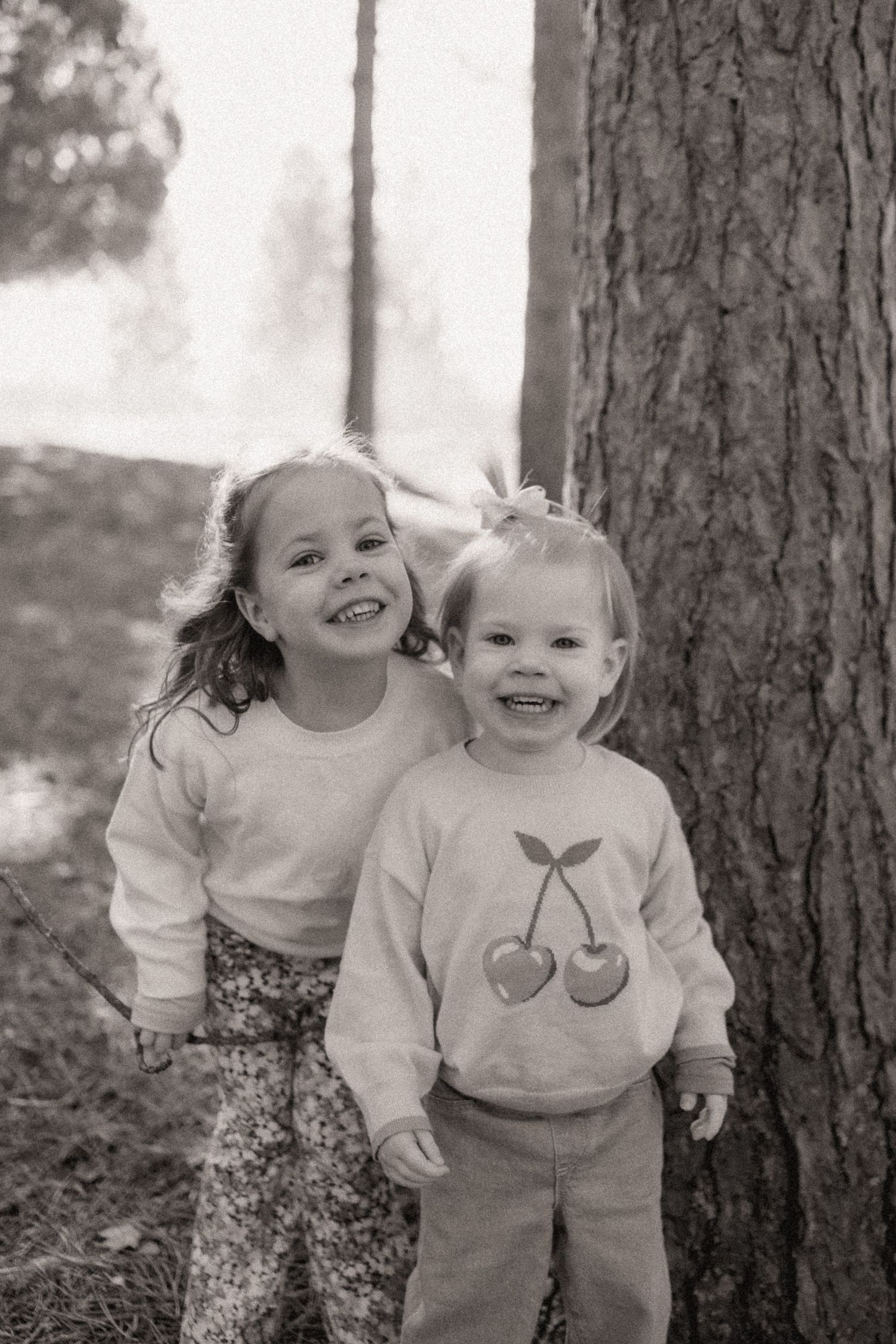 Two young children laughing together in a black and white outdoor portrait series against pine trees.