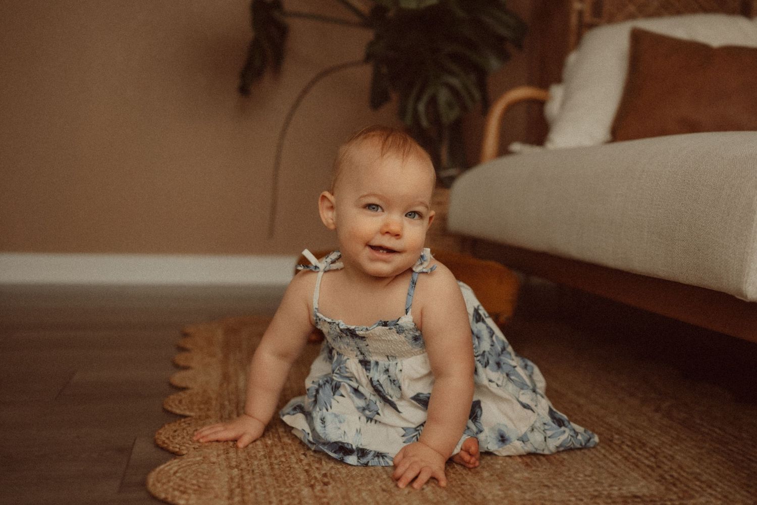 A small child in a floral dress sits smiling on a woven rug near a couch.