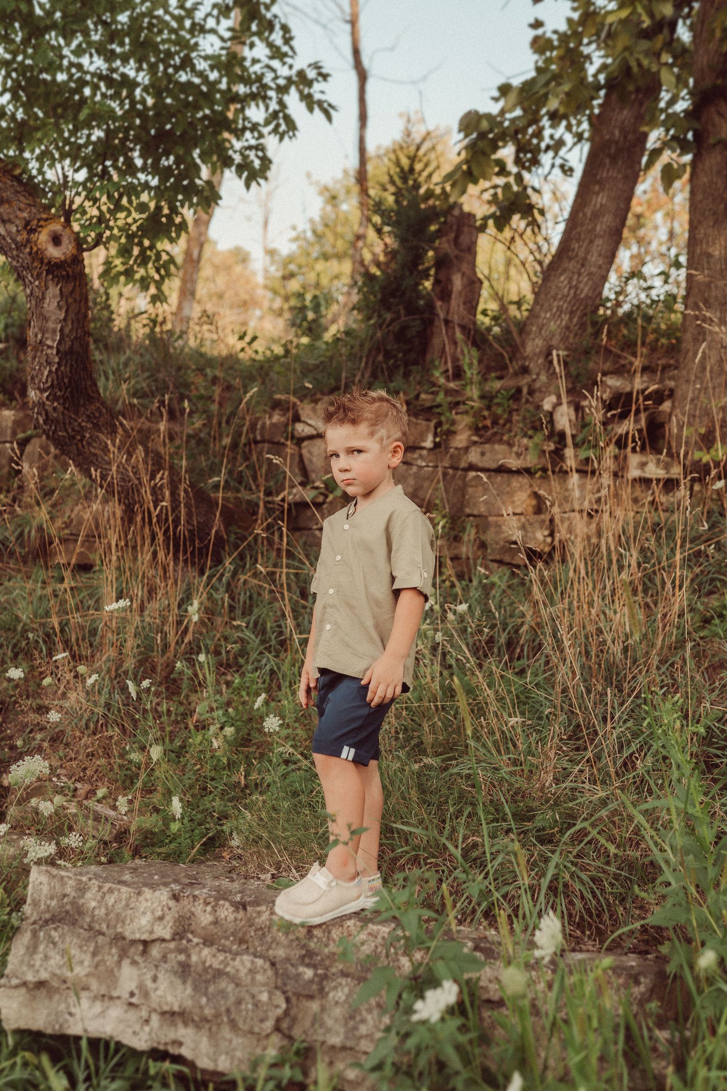 Child in grey shirt and blue shorts stands on stone ledge surrounded by tall grass and trees.
