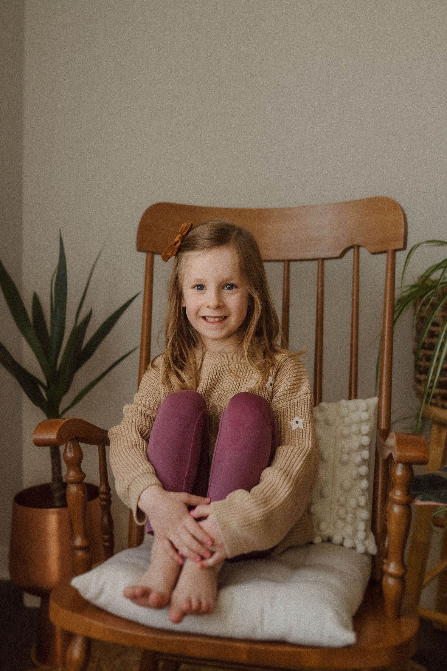 Child in beige sweater and purple pants sits comfortably in wooden rocking chair near plants.