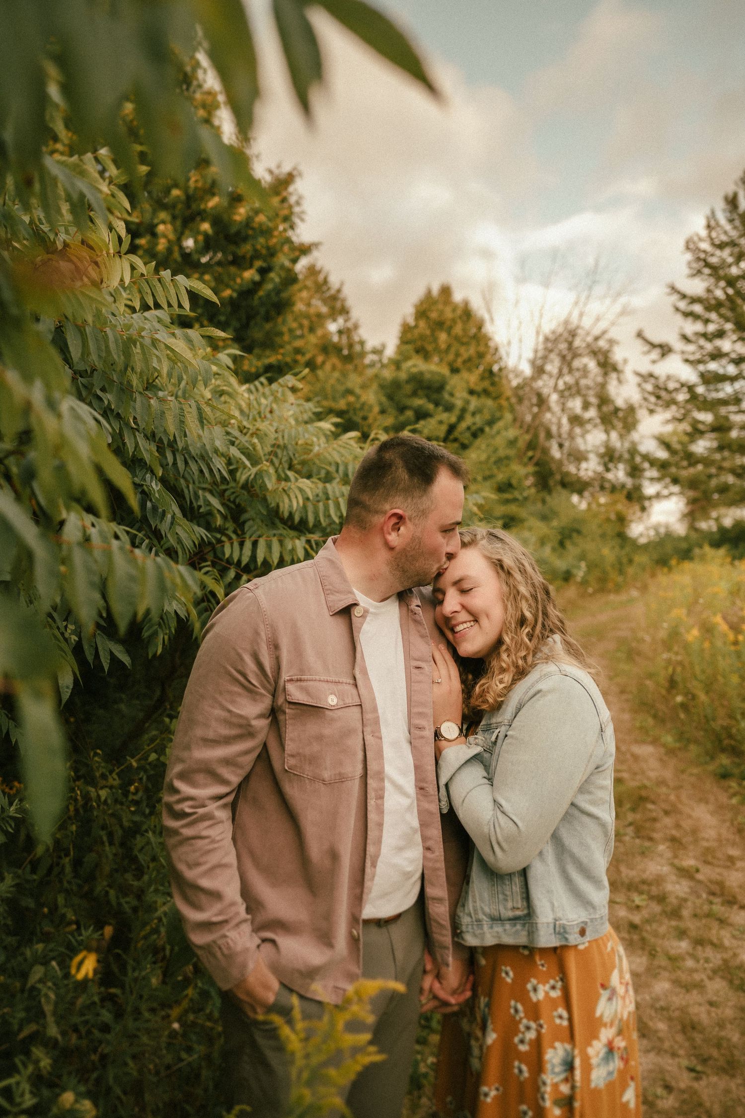 A romantic couple shares an intimate moment on a nature trail surrounded by lush greenery and golden light.