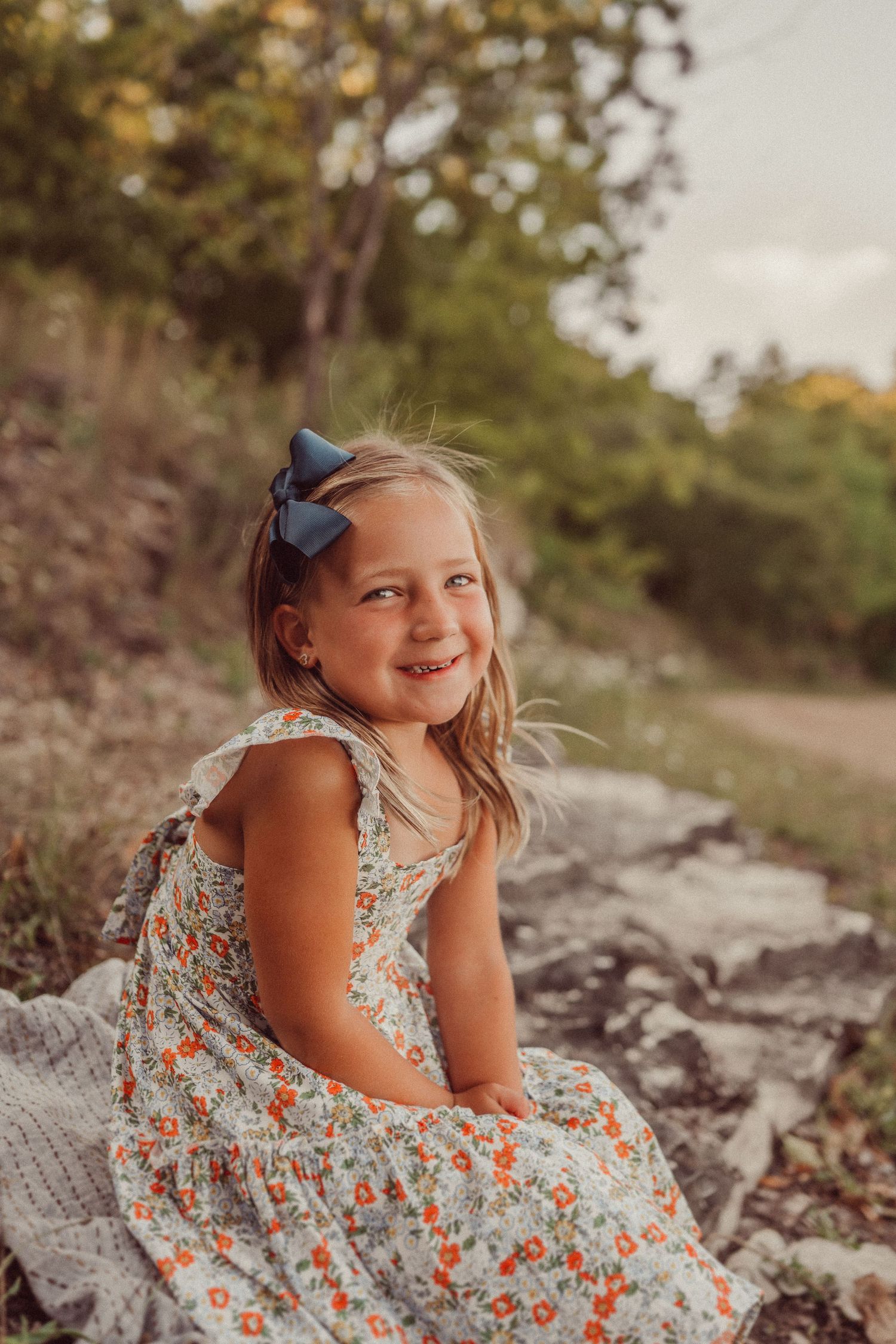 A child in a floral dress and blue bow sits on rocky terrain during golden hour.