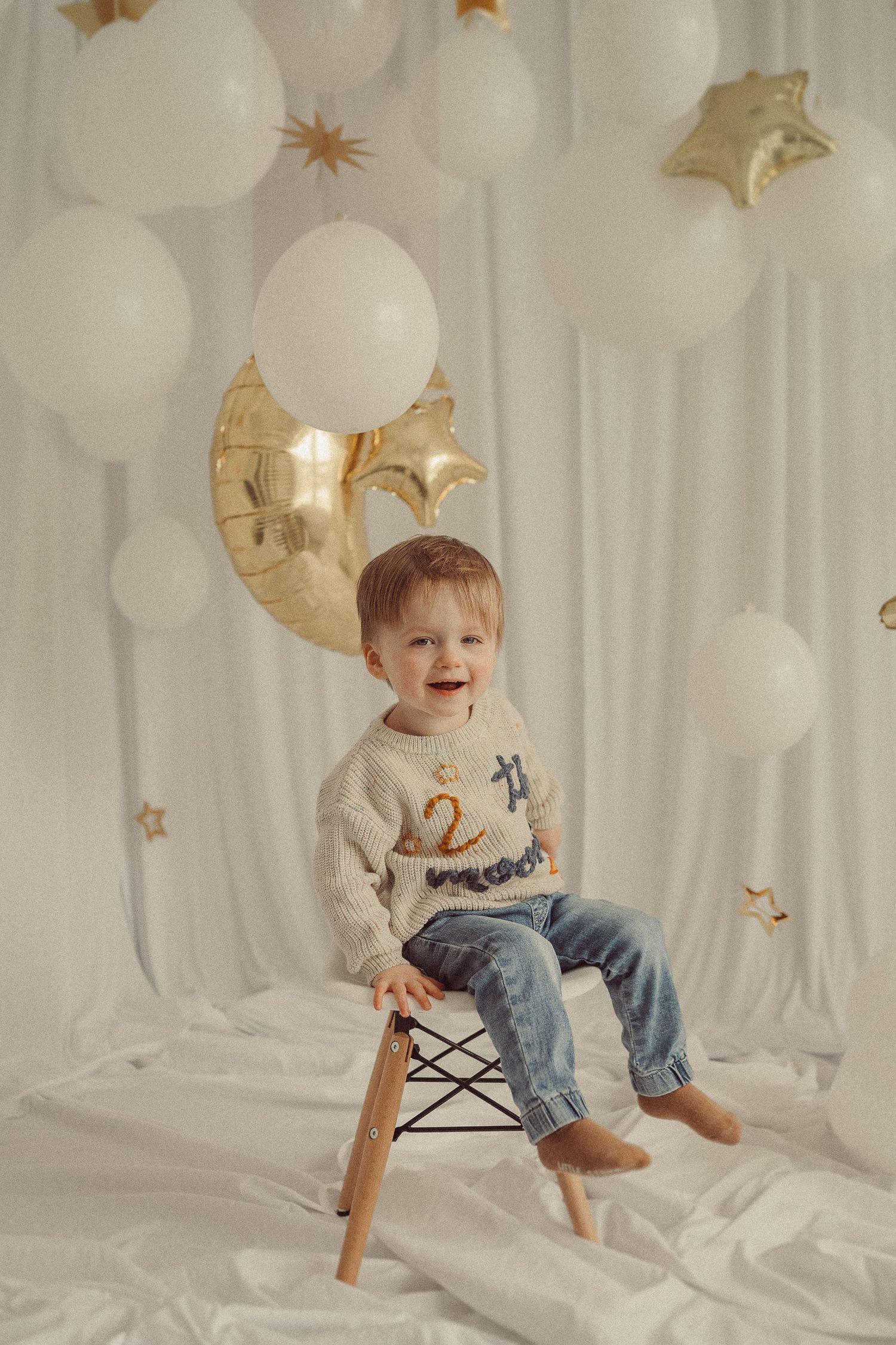 Child sitting on wooden Eames chair against white backdrop with gold stars and balloons.