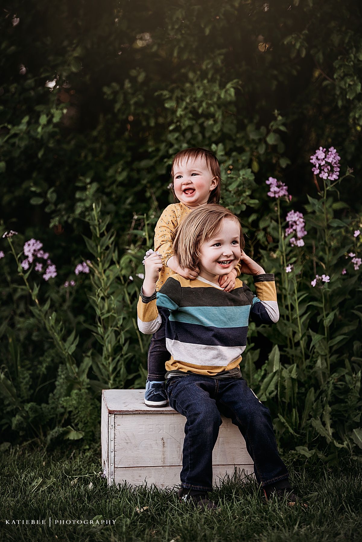 Two children playing on white box with purple wildflowers blooming in background.