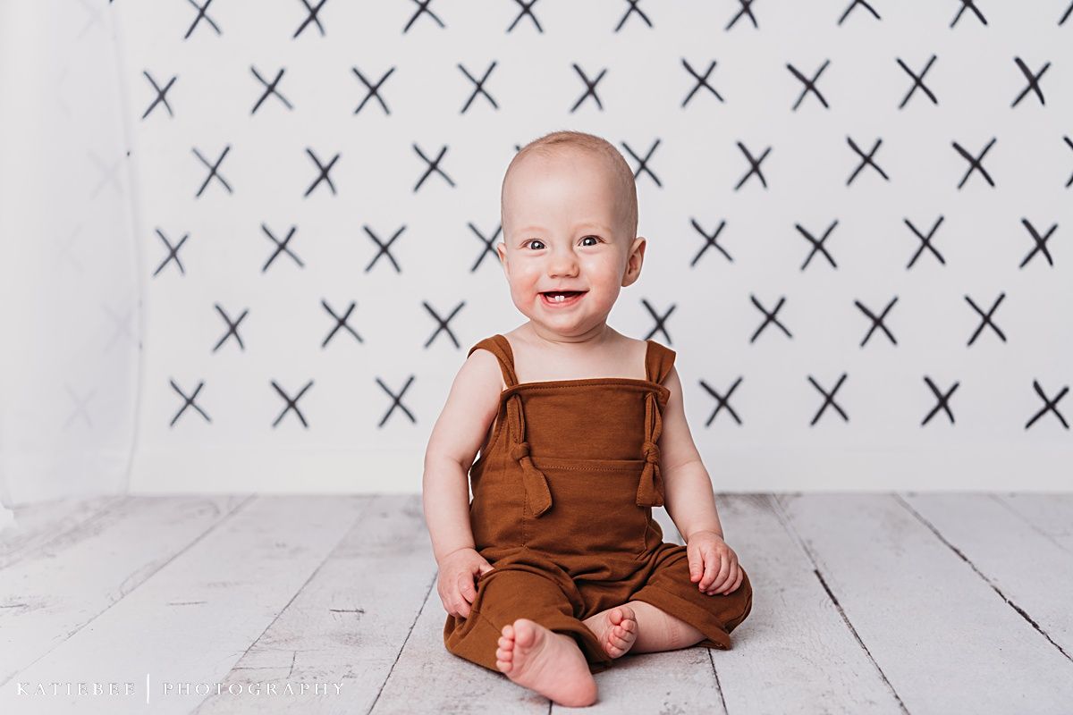 Baby wearing brown overalls sits smiling on white wooden floor with X pattern background.