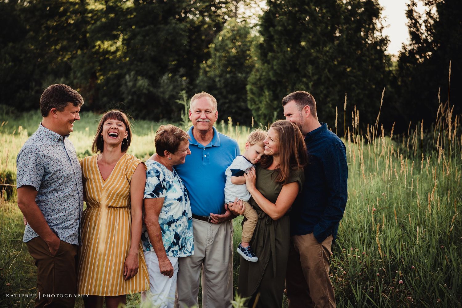 Multi-generational family gathering outdoors in tall grass at sunset.