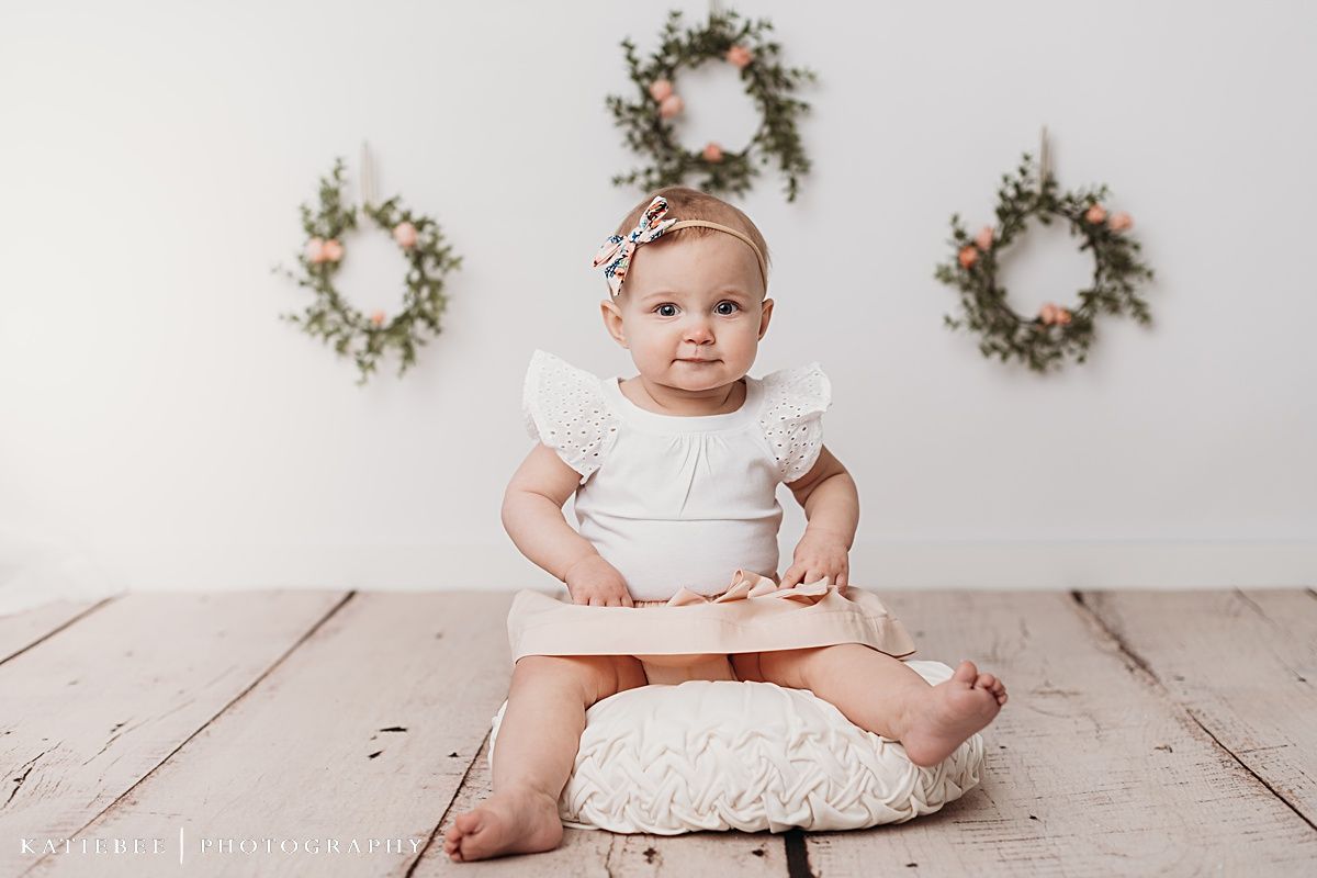 Baby in white dress sitting on cushion against white wall with decorative wreaths.