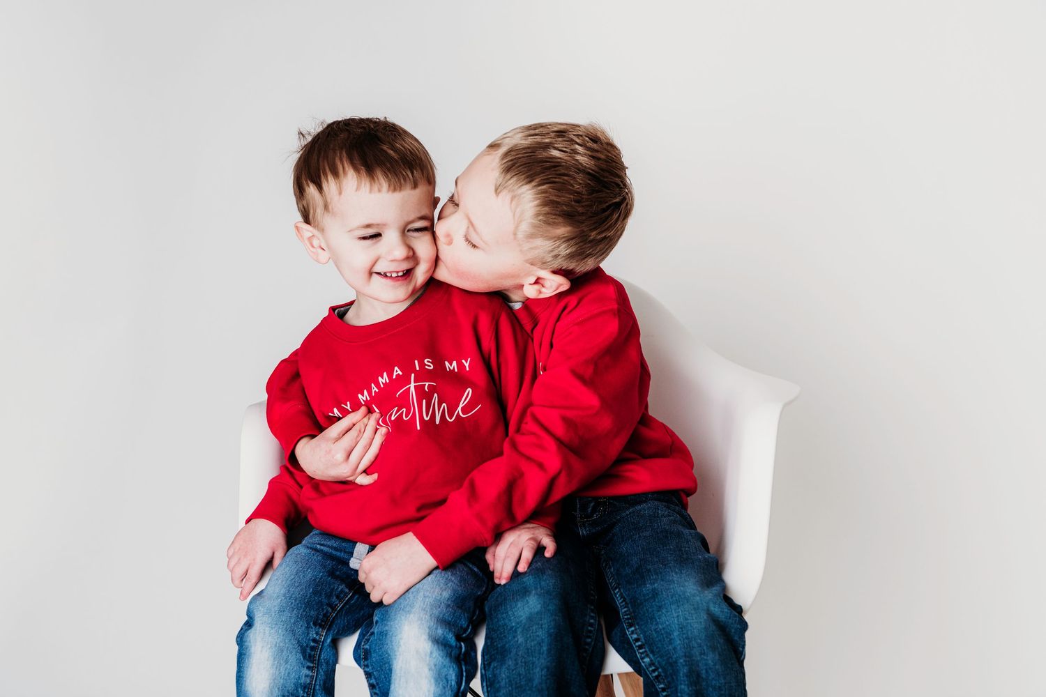 Two young ones in matching red sweaters share a sweet moment while sitting together on a white chair.
