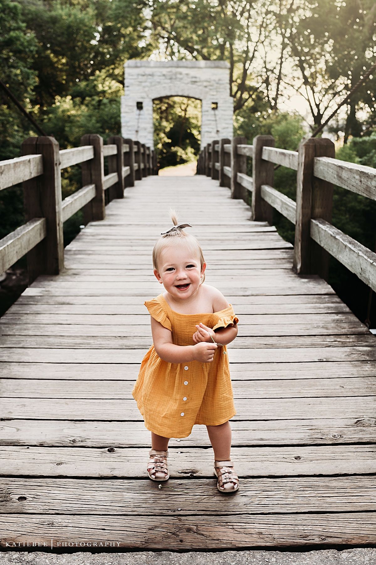 Child in yellow dress laughing on wooden bridge pathway.