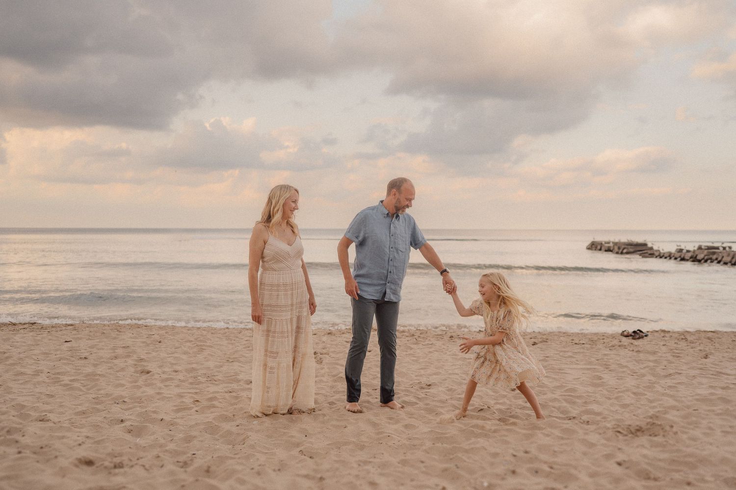 A family holds hands and walks together along a peaceful beach at sunset with cloudy skies above.