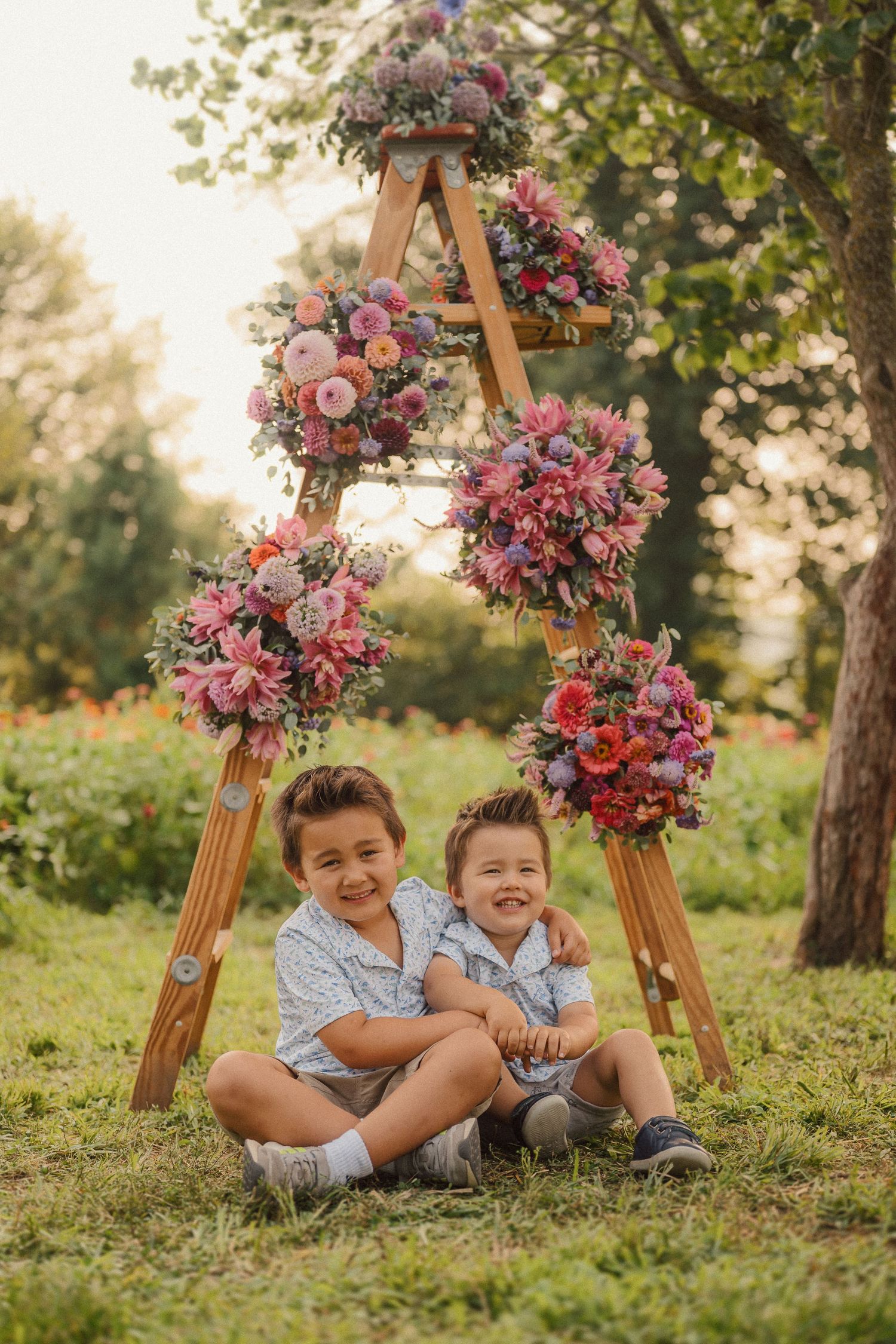 Two joyful children sit under a wooden arch decorated with vibrant pink and red floral arrangements in a garden setting.