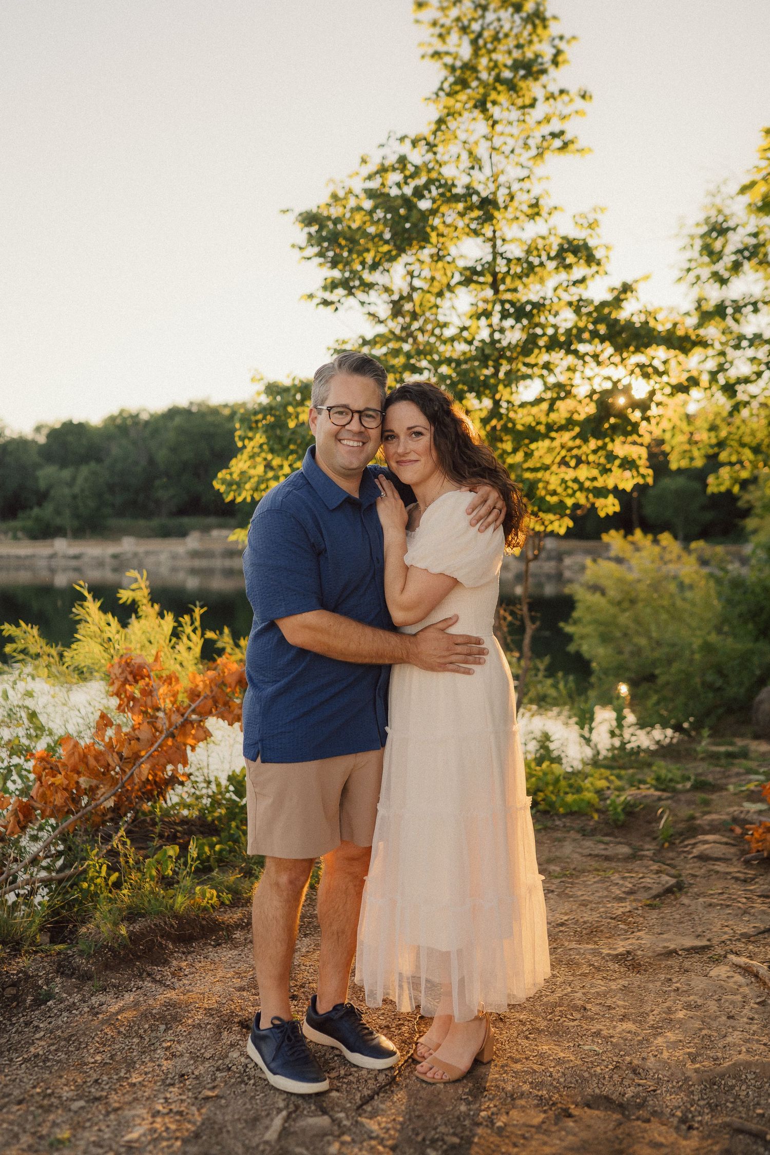 Couple embraces during golden hour photoshoot by lake with autumn foliage in background.