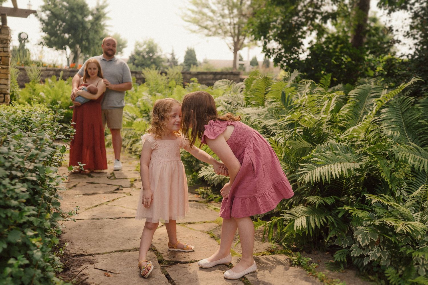 Children in pink and coral dresses explore a lush garden pathway lined with greenery and trees.