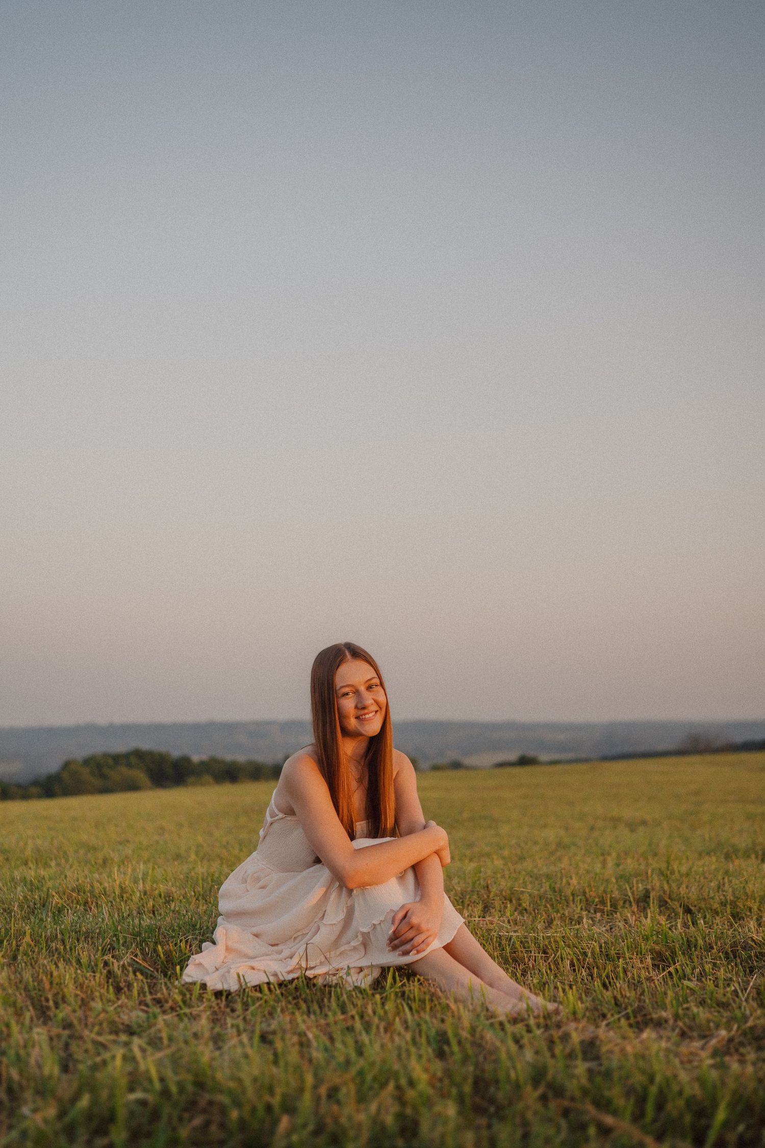 A figure in a white dress sits gracefully on grassy hills during sunset.