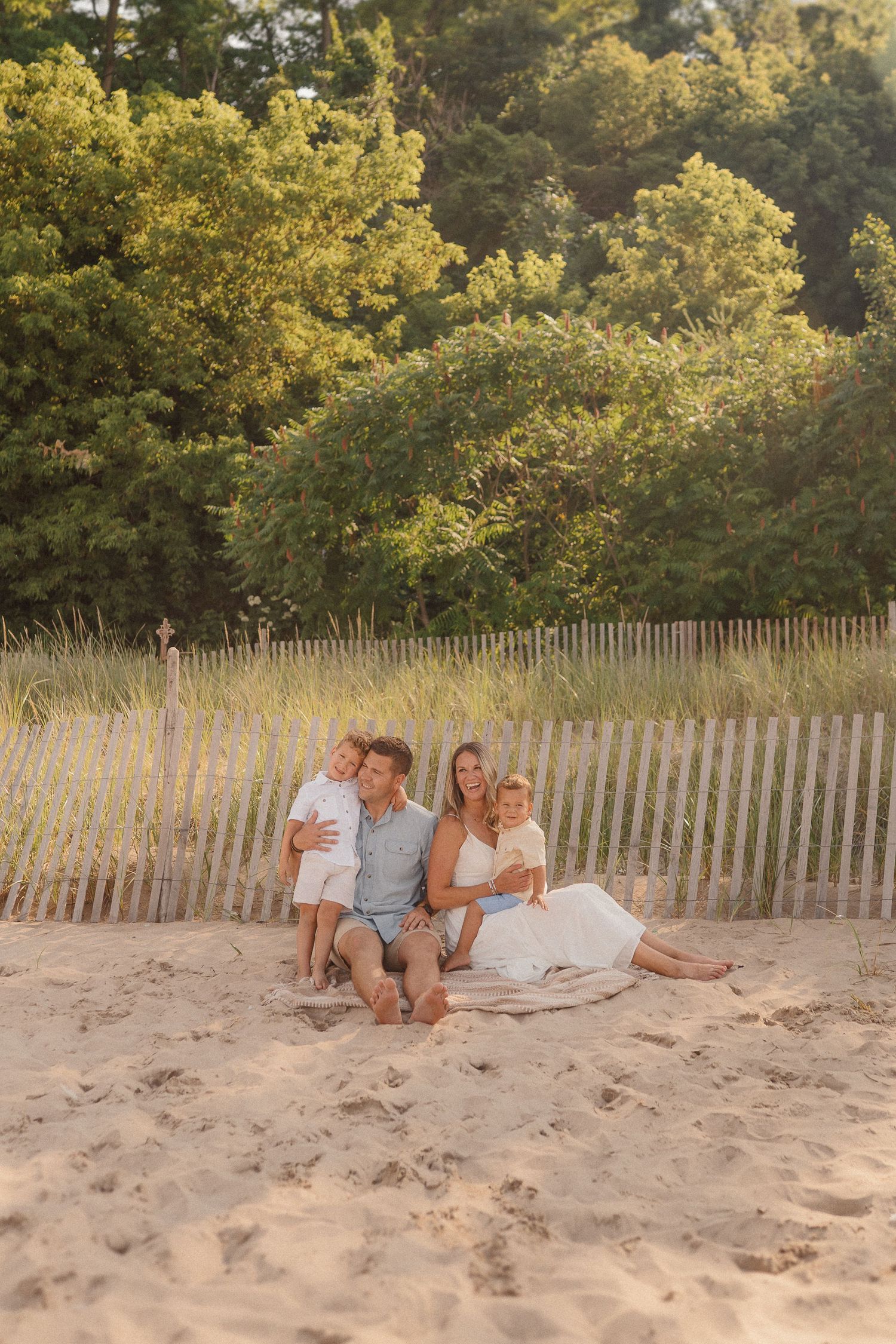 Family relaxing together on sandy beach with wooden fence and golden sunlit trees in background.