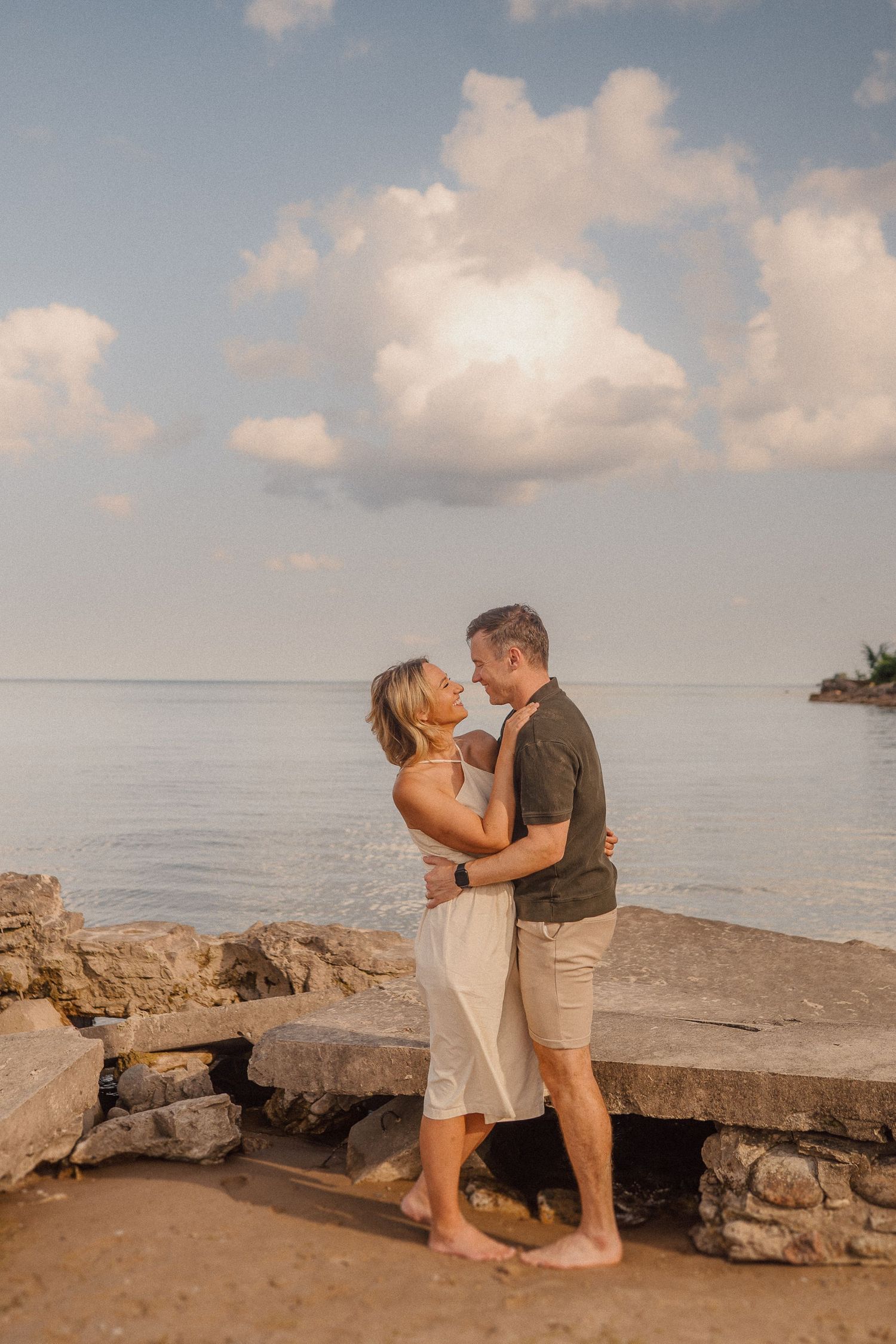 A romantic couple embraces on rocky beach shoreline during golden hour with puffy clouds in background.