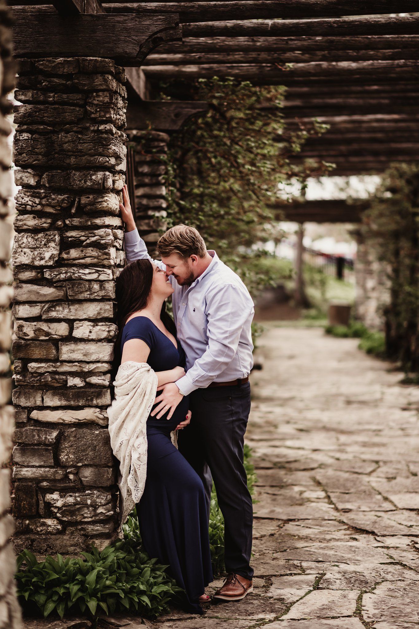 Intimate maternity moment captured against stone pillar in garden setting.
