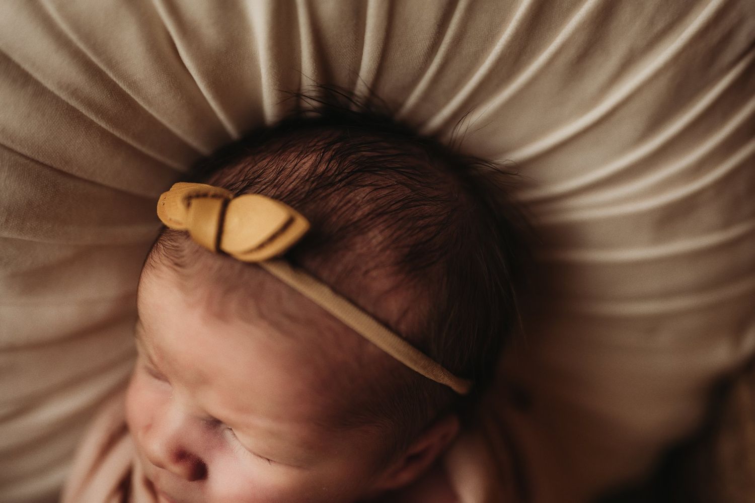 Close-up of sleeping newborn wearing golden bow headband against beige pillow backdrop.
