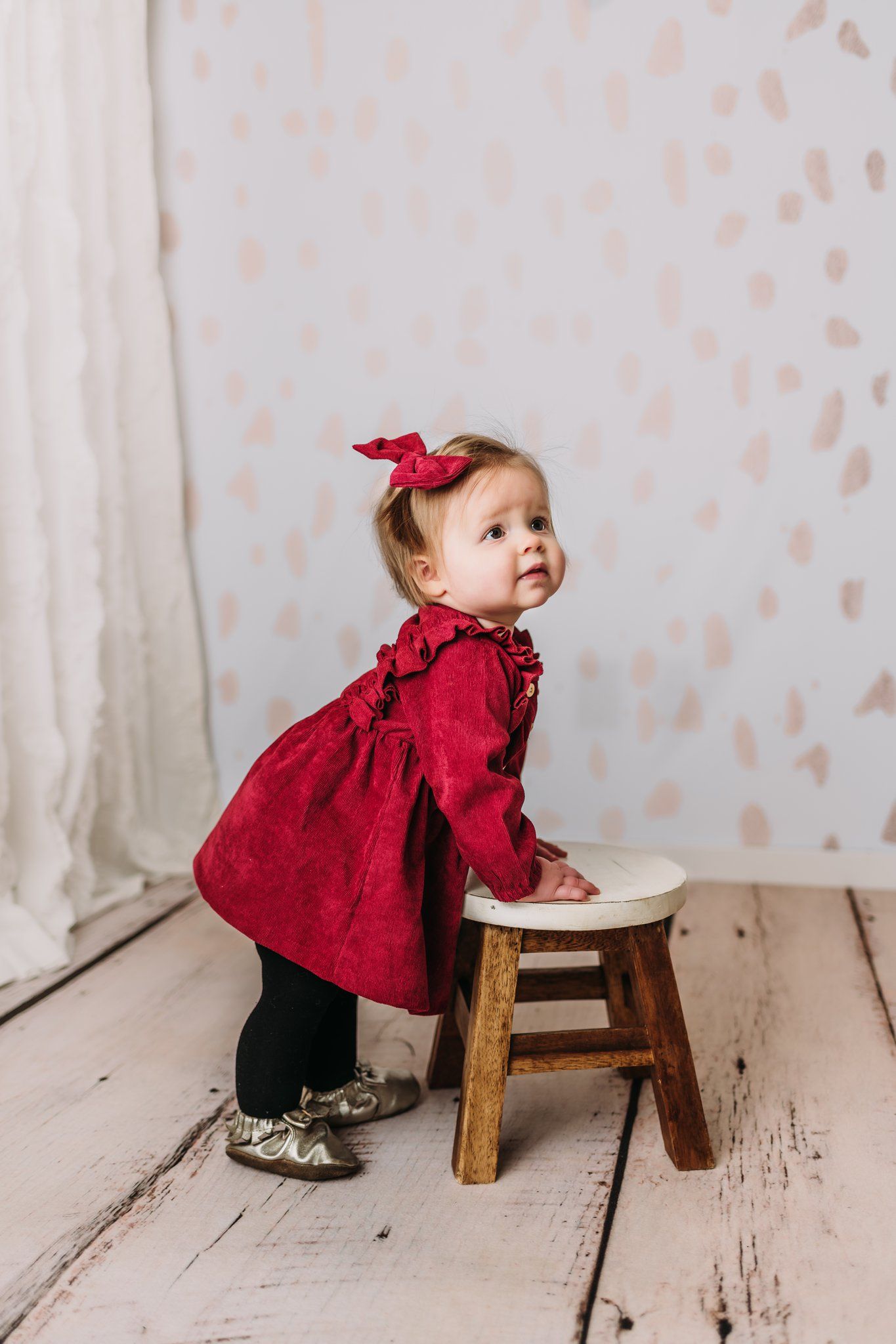Baby in red dress and bow leans against wooden stool on white floor.