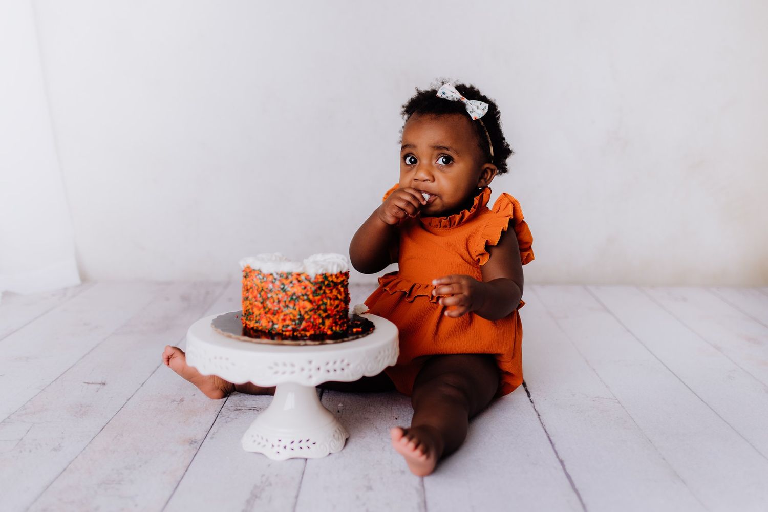 A small child in an orange dress sits next to a cake on a white pedestal.