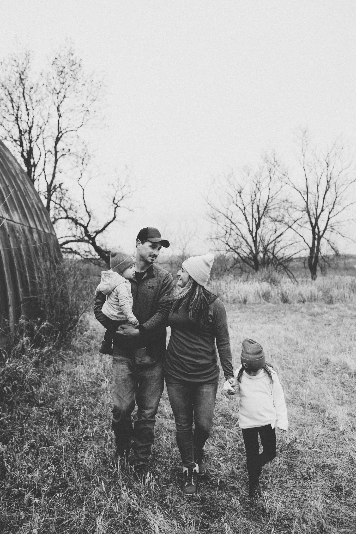 Black and white family photo walking together by rustic barn in winter.