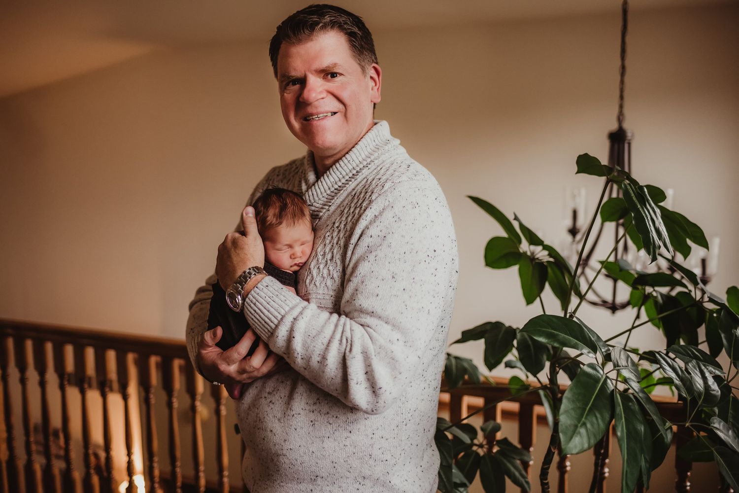 Person in cream sweater holding infant near indoor houseplant and wooden staircase.