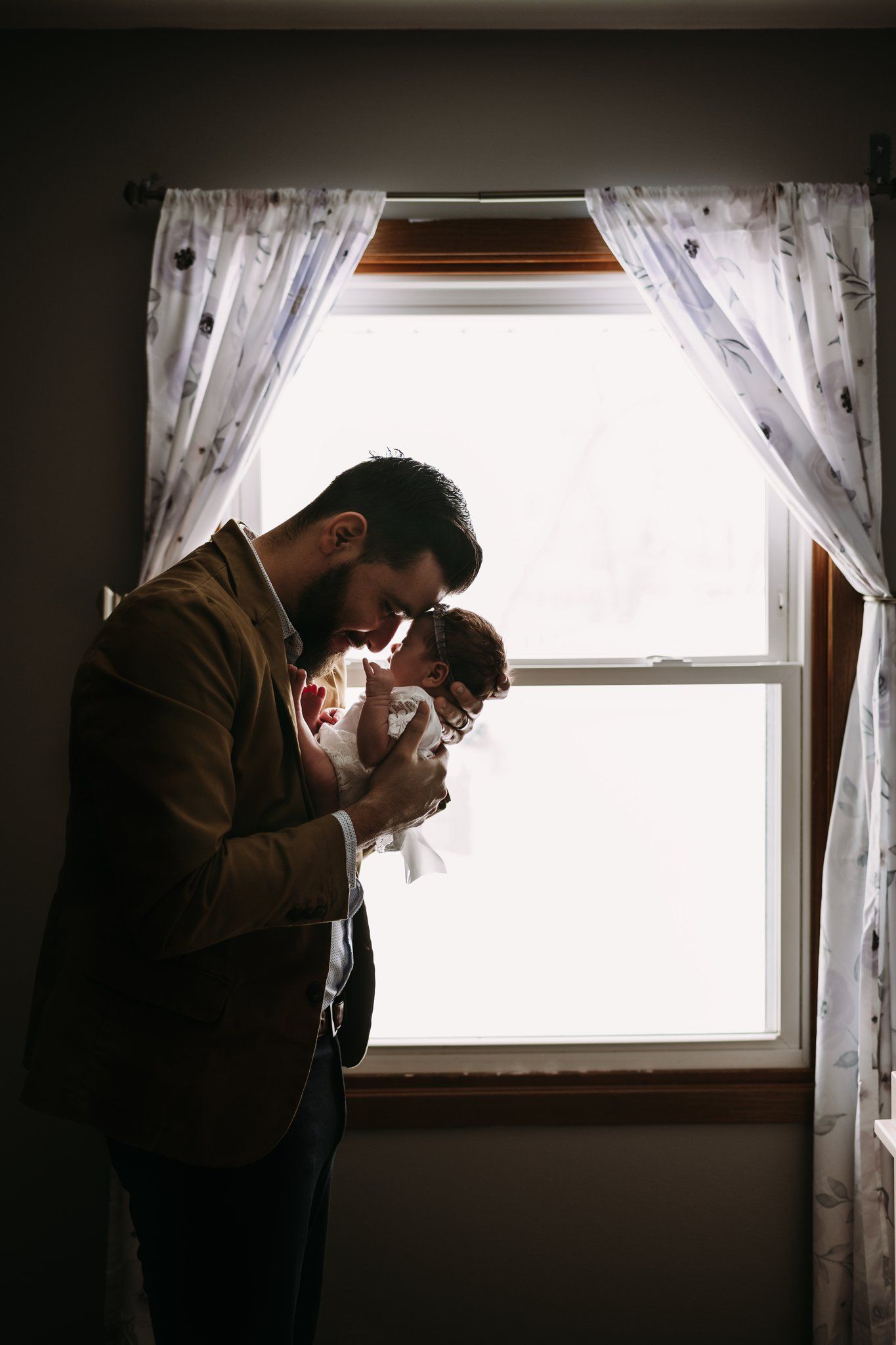Silhouetted figure in brown jacket holds infant by window with white curtains.