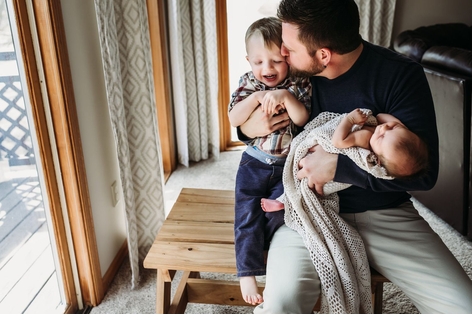 Parent skillfully holds both a toddler and swaddled baby near sunlit windows with wooden bench.