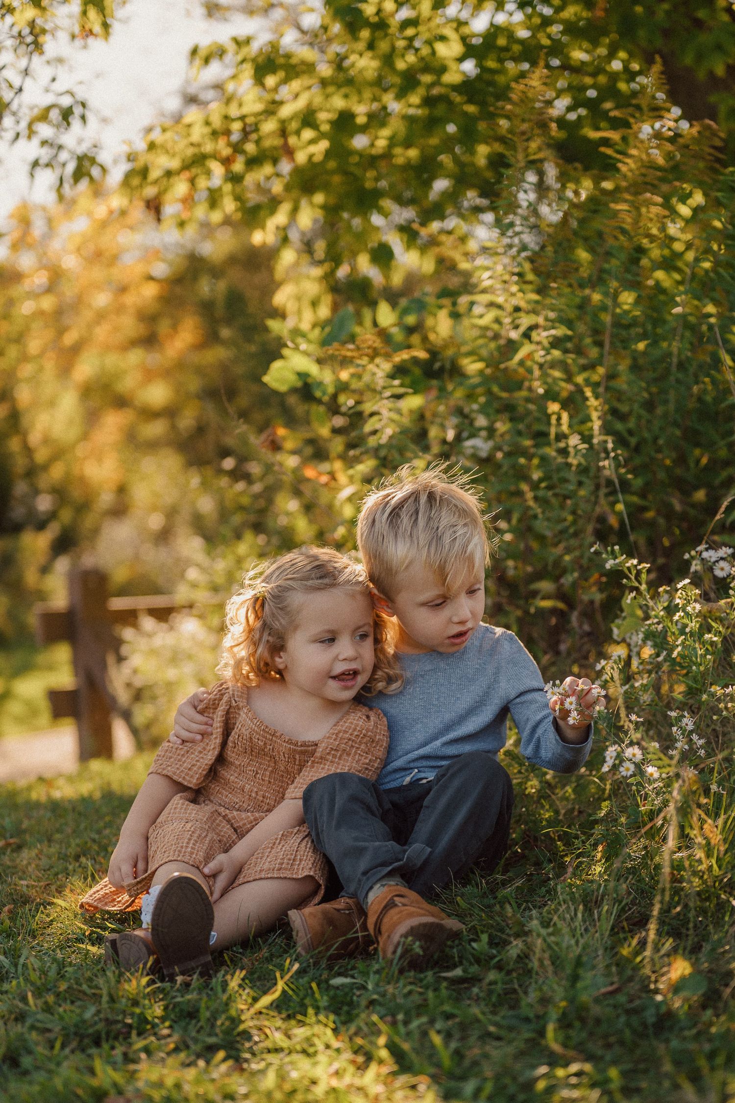 Two children sitting together on grass under autumn trees during golden hour.