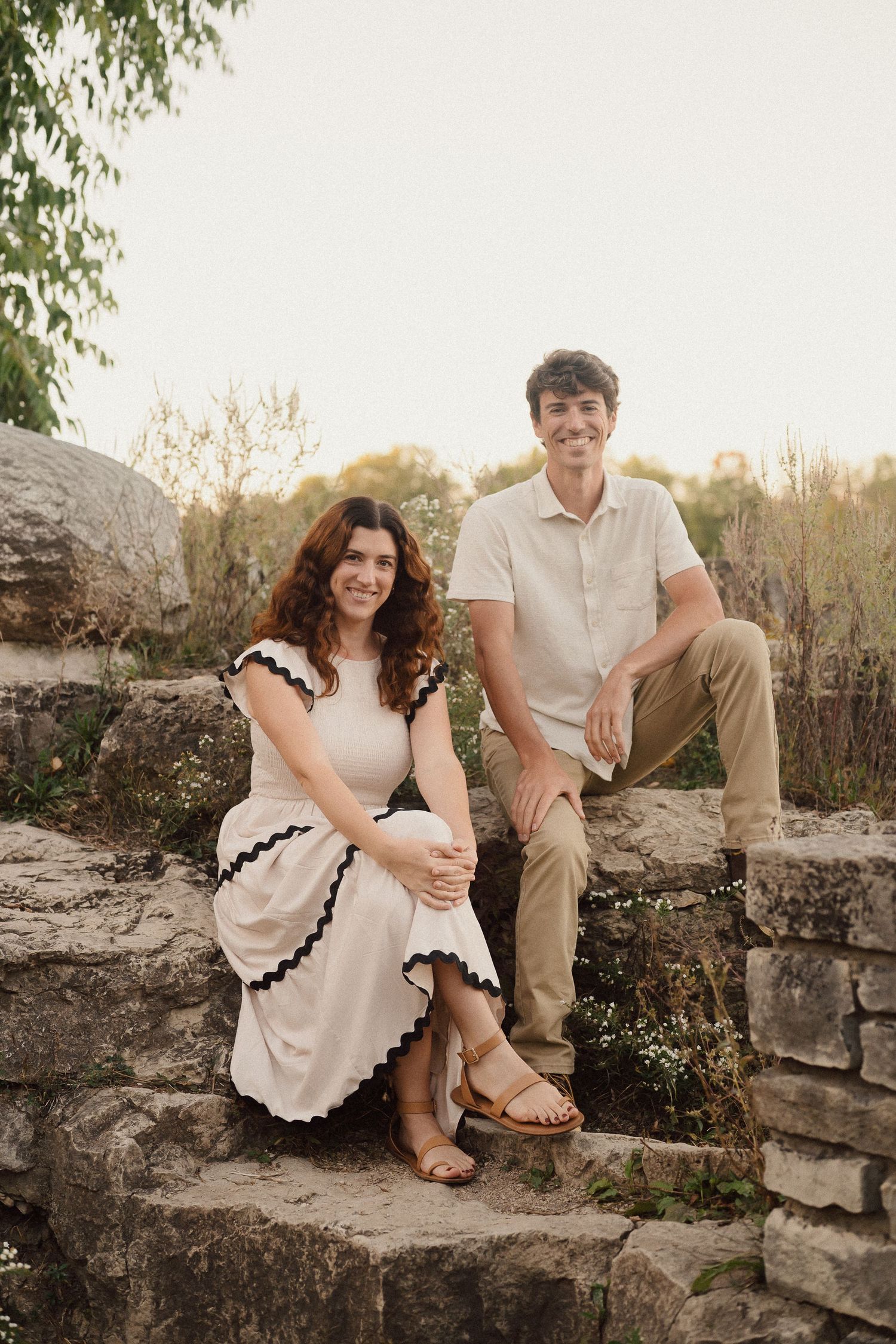 A couple sits together on a rustic stone wall during an outdoor photo session at sunset.