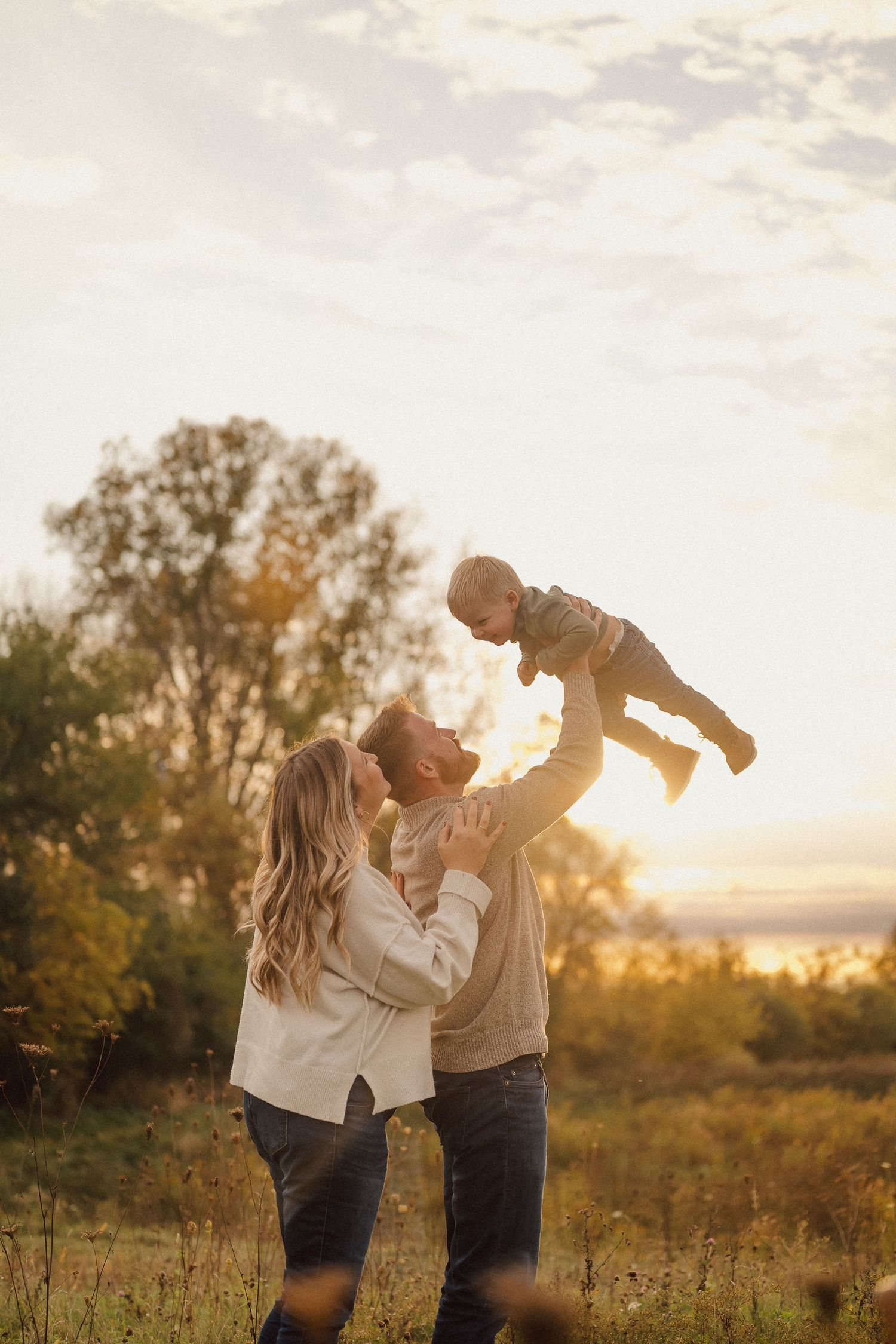 A couple lifts their child in the air against a sunset backdrop in a field.