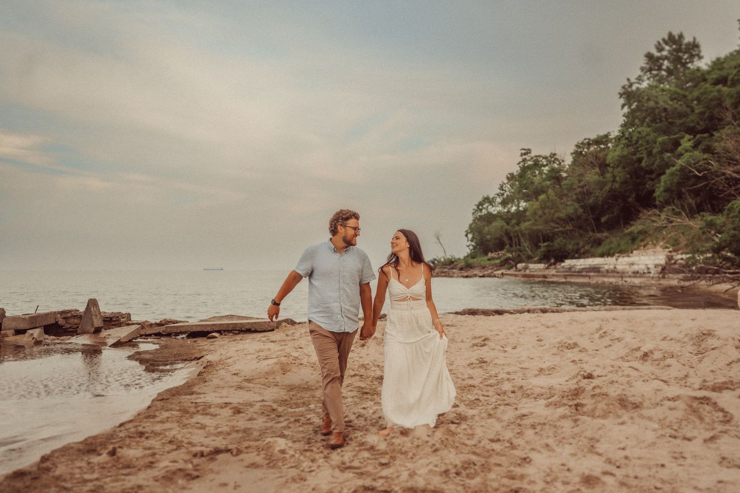 Couple walking hand in hand on sandy beach at sunset wearing casual attire.