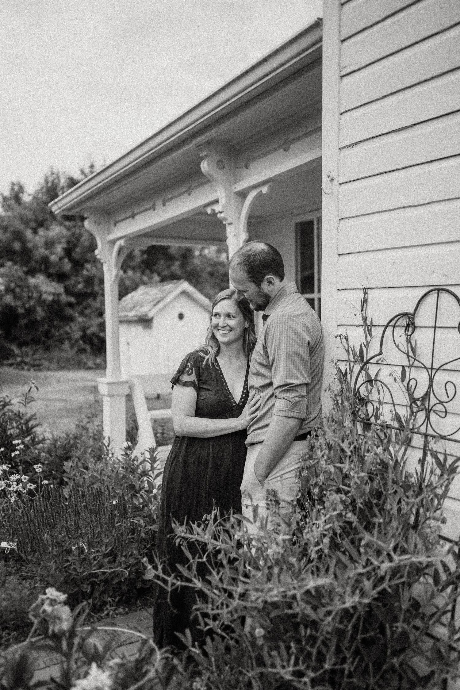 Black and white portrait of couple by white farmhouse with garden.